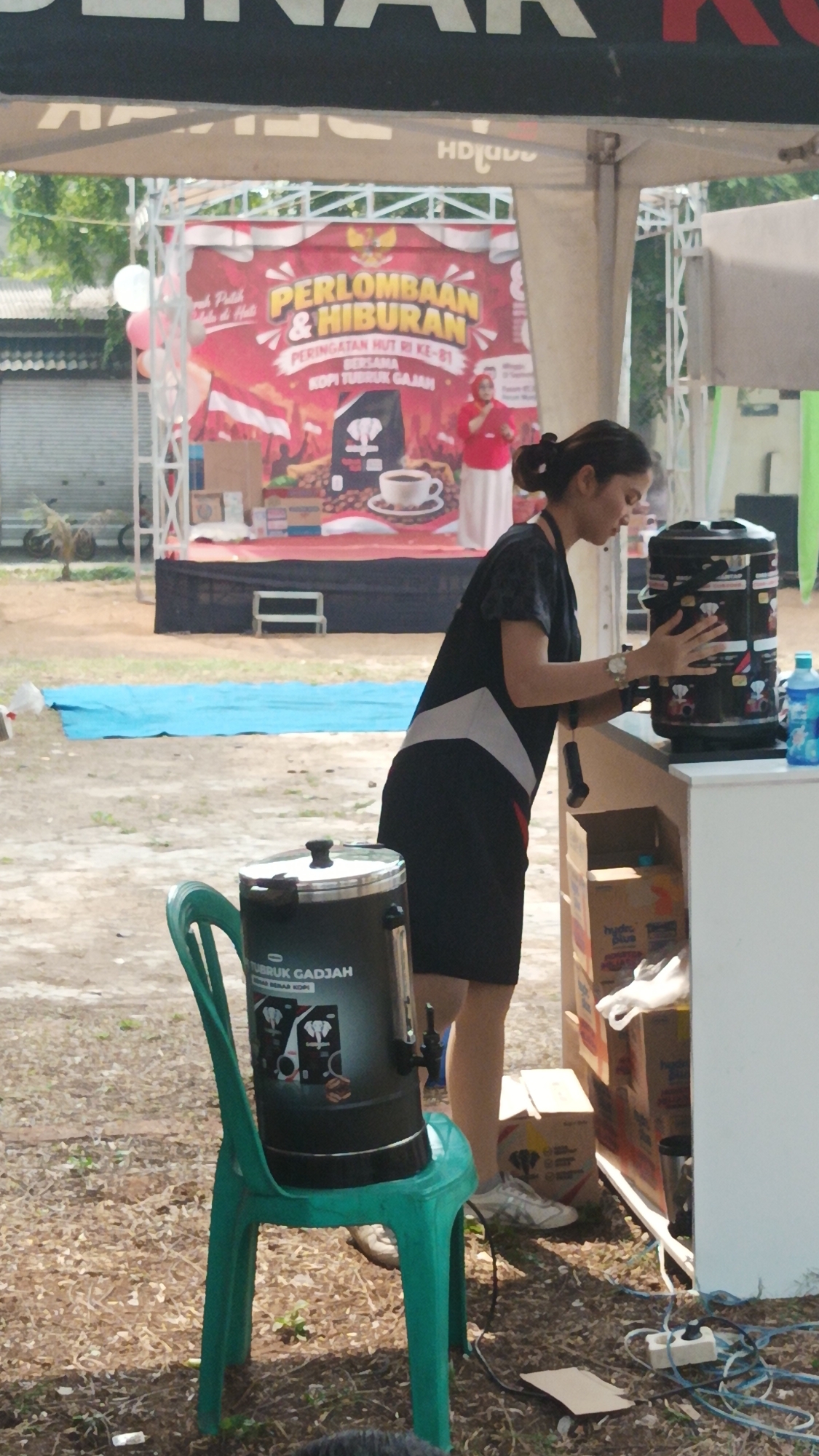 A woman is operating a commercial coffee dispenser with a black and silver exterior, adorned with stickers featuring elephants and coffee beans, at an outdoor event. She is wearing a dark dress with white and red accents and has her hair pulled back. To her left, a large black coffee urn with a prominent label "KOPI TUBRUK GAJAH" sits on a green plastic chair. In the background, a red banner with white and yellow text, including "PERLOMBAAN & HIBURAN" and "PERINGATAN HUT RI KE-81", is displayed. The scene appears to be a local festival or market in Munjul, Indonesia, with tents, stalls, and a crowd visible. The ground is covered in dry leaves and dirt, suggesting an outdoor setting. Several cardboard boxes are stacked near the coffee dispenser.