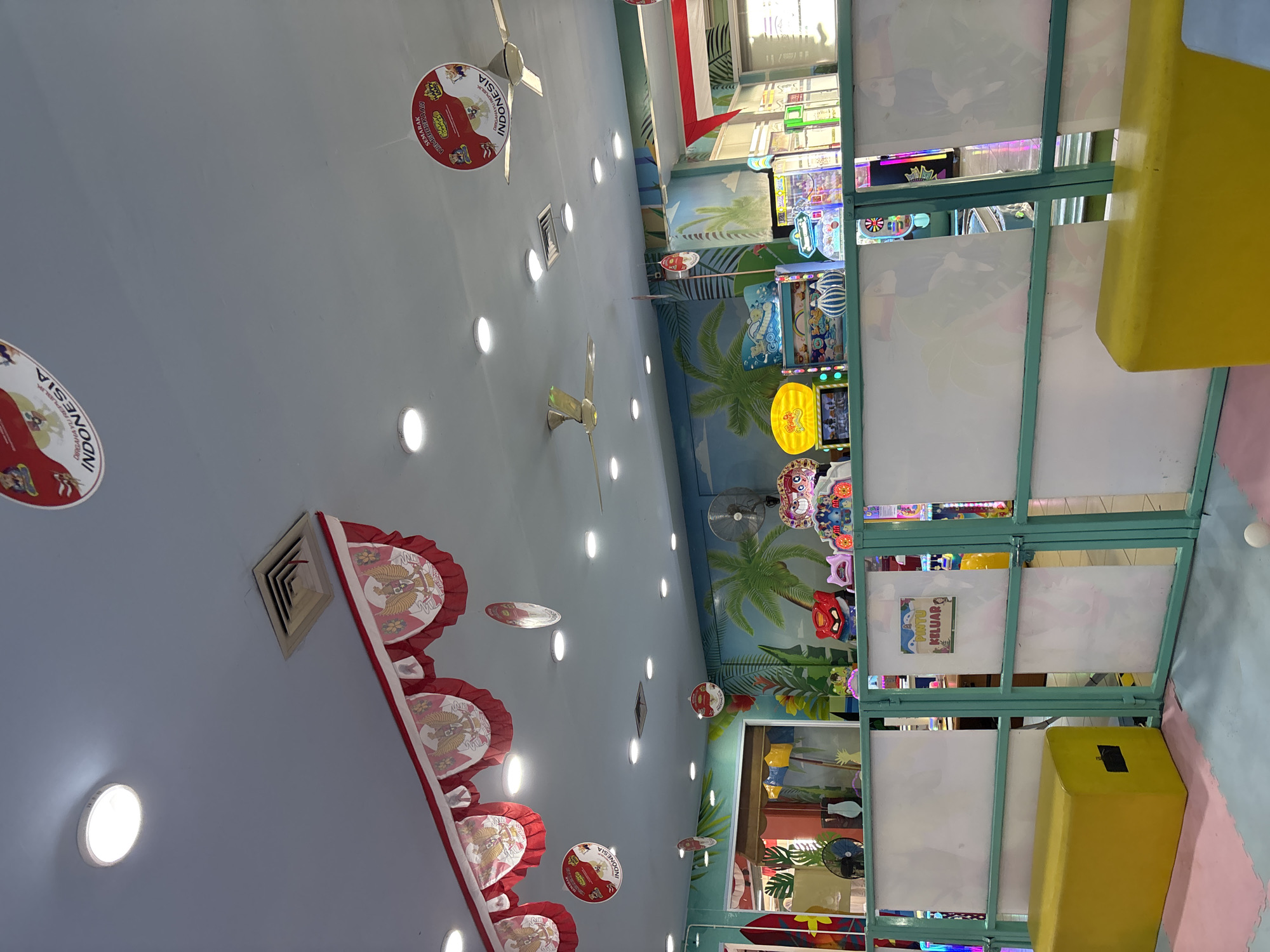 The image shows an indoor amusement or arcade area decorated with Indonesian national symbols. Red and white banners featuring the Garuda Pancasila, the national emblem of Indonesia, are hung along the ceiling. Circular decorations with the text "Dirgahayu Republik Indonesia" are also visible. The environment appears to be a children's play area with colorful murals depicting palm trees and a tropical scene on the walls. Several arcade machines are present, some featuring animated characters and bright lights. A yellow bench is placed in the foreground. The overall impression is of a festive and lively space, likely celebrating Indonesian Independence Day. The location is identified as Liangjulang, Indonesia.