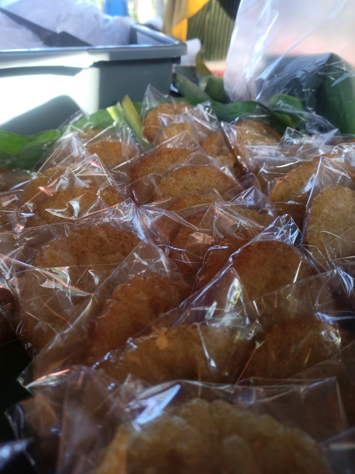 The image shows a close-up view of several small, golden-brown, circular snacks, each individually wrapped in clear plastic. The snacks appear to be deep-fried and have a slightly uneven, textured surface. They are arranged in multiple rows, filling the foreground and extending into the background. Some green leaves are visible interspersed among the snacks. In the background, out of focus, is a grey plastic container and what appears to be a white plastic bag, suggesting a market or food stall setting. The lighting suggests it is daytime, with a bright, diffused light source coming from the left, casting some shadows and highlights on the plastic packaging and the snacks. The overall scene is likely from Karungan, Indonesia, given the context provided.
