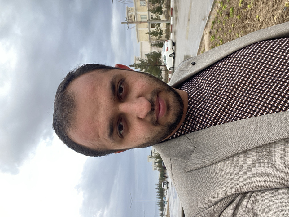 A man poses in front of a drab, overcast sky. He has dark hair, a beard, and brown eyes. He wears a patterned shirt and a grey blazer. Behind him, there's a street with wet pavement, a few buildings, and a car. The scene suggests an urban or suburban setting on a cloudy, possibly rainy, day.