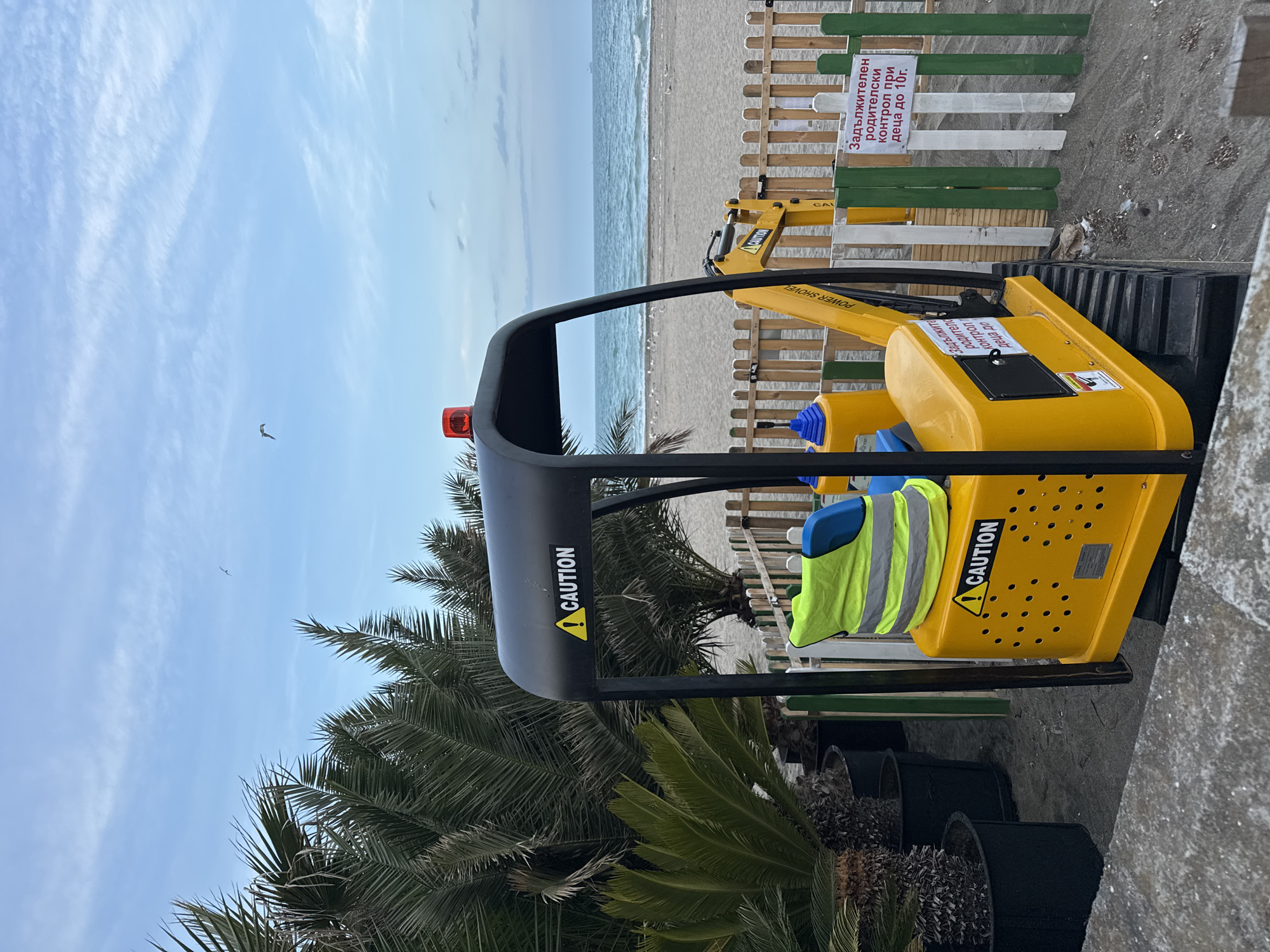 The image captures a beach scene in Burgas, Bulgaria, showcasing a yellow toy excavator positioned on a concrete area, with palm trees providing a partially obscured view of the beach and sea beyond. The excavator has a black overhead structure marked with the word "CAUTION" and a reflective vest draped over the seat. A wooden fence, painted with alternating sections of white and green, separates the excavator from the sandy beach. On the fence, a sign in Bulgarian reads "Задължителен родителски контрол при деца до 10г.", meaning "Mandatory parental control for children up to 10 years old." The sky is partly cloudy. The overall atmosphere is calm and suggestive of a recreational beach setting.