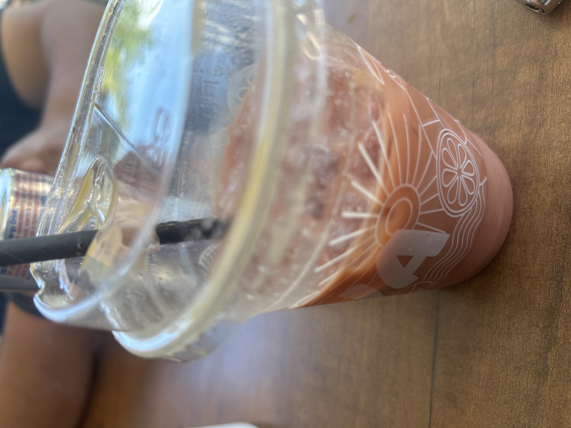 A clear plastic cup with a straw is filled with an orange beverage and ice. The cup has a white design of a sun and flower on it. It sits on a brown wooden table. In the background, a blurry person is visible, and a can of energy drink is also present. The scene is set outdoors, possibly in Osikovitsa, Bulgaria, given the context provided, though the image itself does not contain explicit location markers.