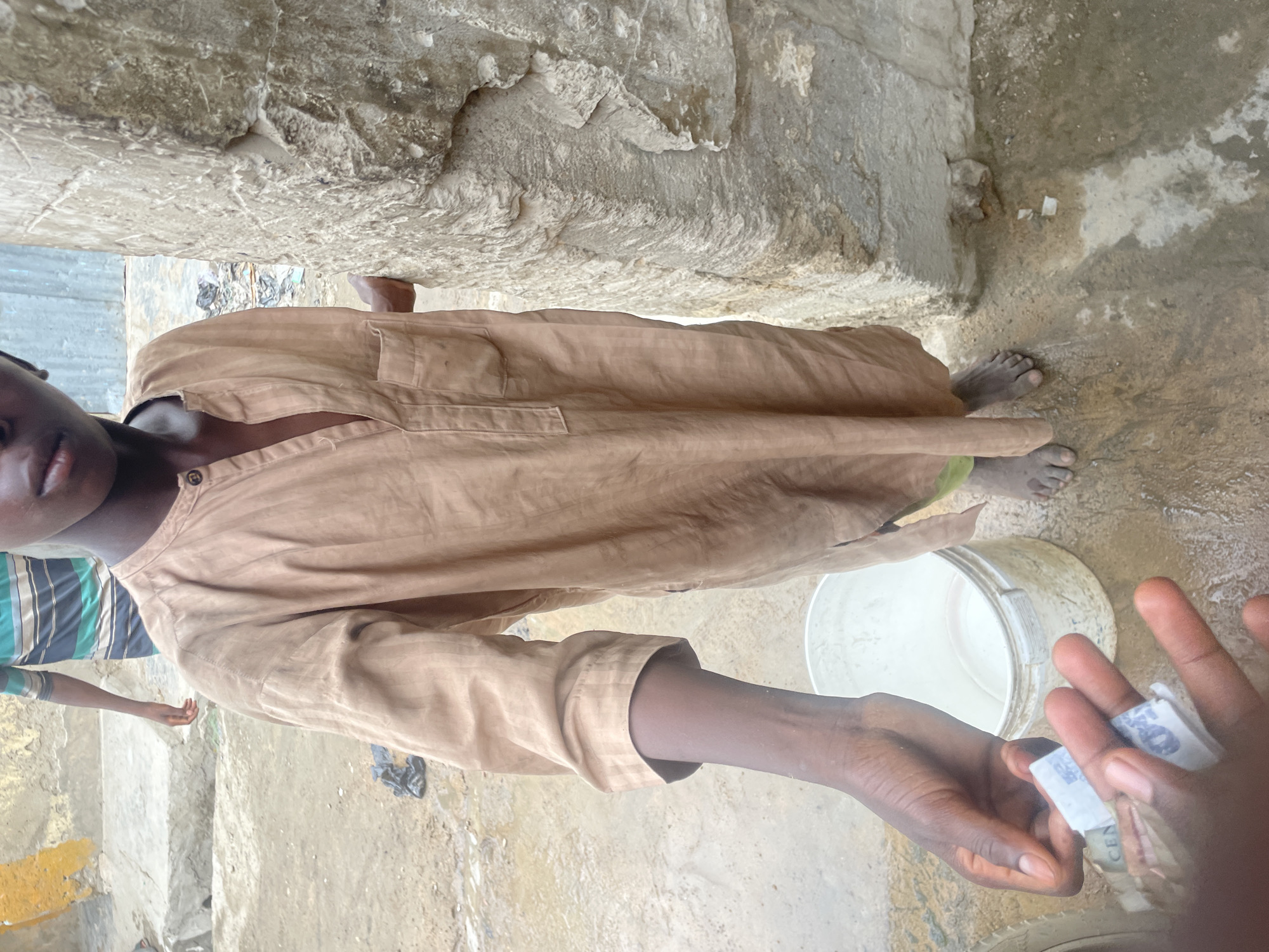 A street scene in an unidentified city shows a young person in a brown tunic. Another person, visible only by their arm, is handing them currency. A bucket and some wet ground are visible, along with some concrete walls and steps.