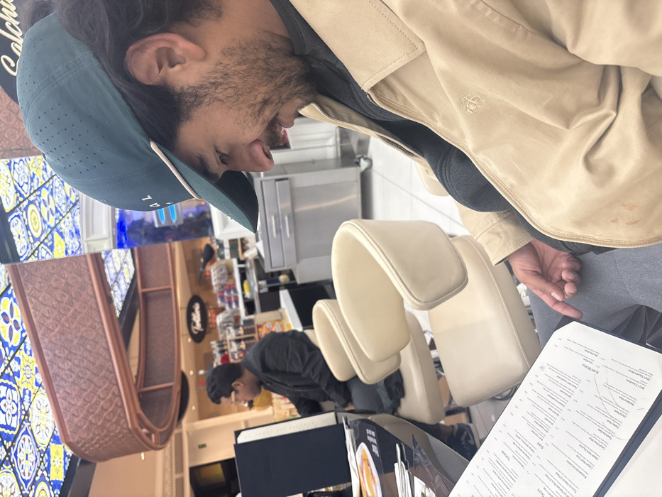 A man in a tan jacket and teal cap peruses a menu in a brightly lit restaurant. Beige chairs are visible. In the background, another man in a black jacket is standing near a counter. A decorative tiled wall and a textured archway add to the restaurant's aesthetic. The photo appears to be taken from an elevated angle, possibly from someone seated at a nearby table.