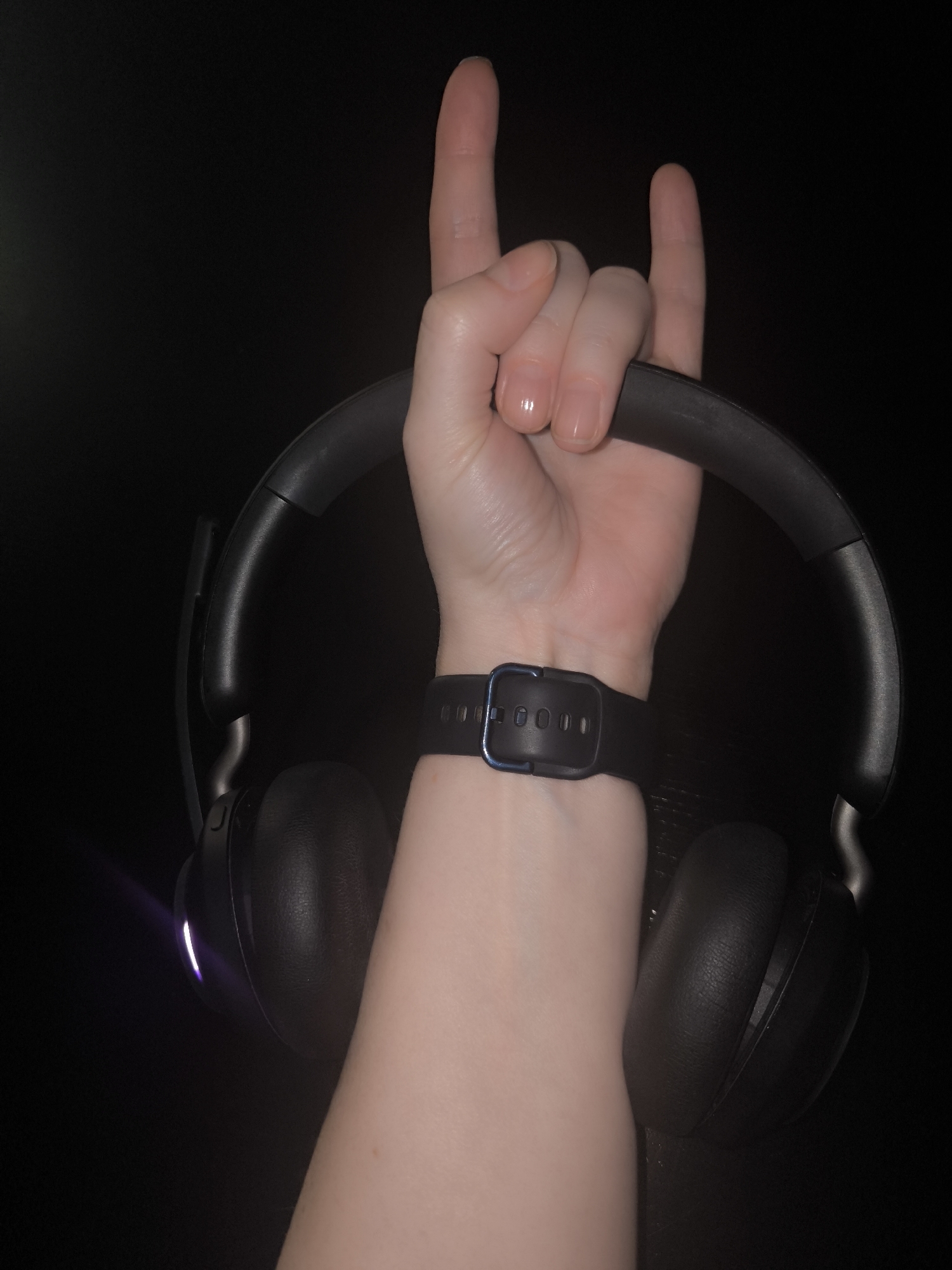 A right forearm and hand are shown against a black background, holding black headphones. The hand is positioned with the index and pinky fingers extended upwards, forming a "rock on" or "devil horns" gesture. A black wristwatch is visible on the wrist. The scene's location is Unknown, Unknown.