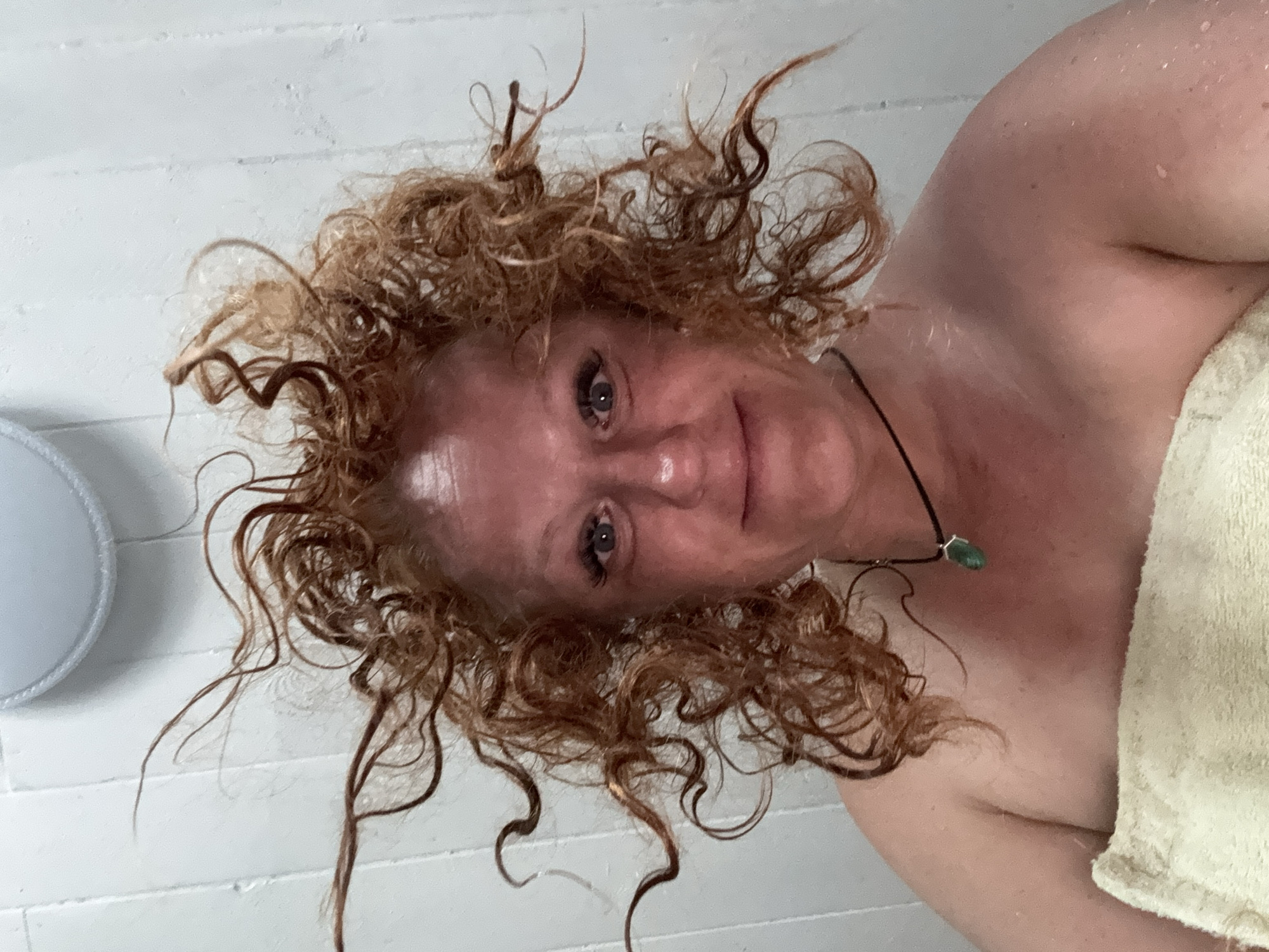 An upward-angled selfie captures a woman, likely in a bathroom or shower setting, with her head slightly tilted and a gentle, contented smile directed at the camera. Her reddish-brown, curly hair is visibly wet and disheveled, with numerous damp strands framing her face. Water droplets are apparent on her skin, particularly across her forehead, shoulders, and upper chest, indicating she has recently showered or bathed. She has blue or grey eyes. Around her neck, she wears a simple dark cord necklace adorned with a small, green, possibly hexagonal, pendant. A light-colored, likely cream or light yellow, towel is draped around her body, covering her from the shoulders down. The background features a white wall with horizontal lines, suggesting tiling or paneling. A blurred, light-toned, circular object is partially visible on the left side of the background. There is no visible text in the image. While the provided context indicates the location is Chaikovsky, Russia, the image itself does not contain specific visual cues to confirm this.