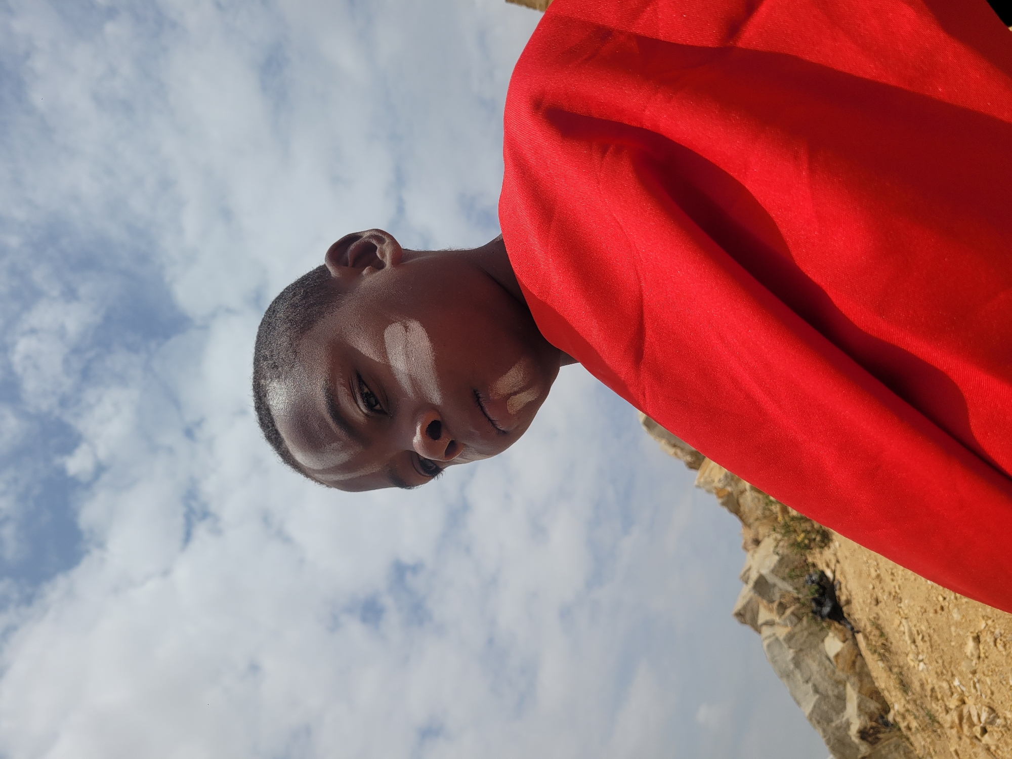 A low-angle, slightly rotated close-up shot features a Black male wearing what appears to be a red cloth. He has short hair, and there are visible white markings on his face. Behind him, the sky is filled with scattered clouds. The foreground is a rocky, dirt terrain. This image was taken in the city of Barakin Akawo, Nigeria.