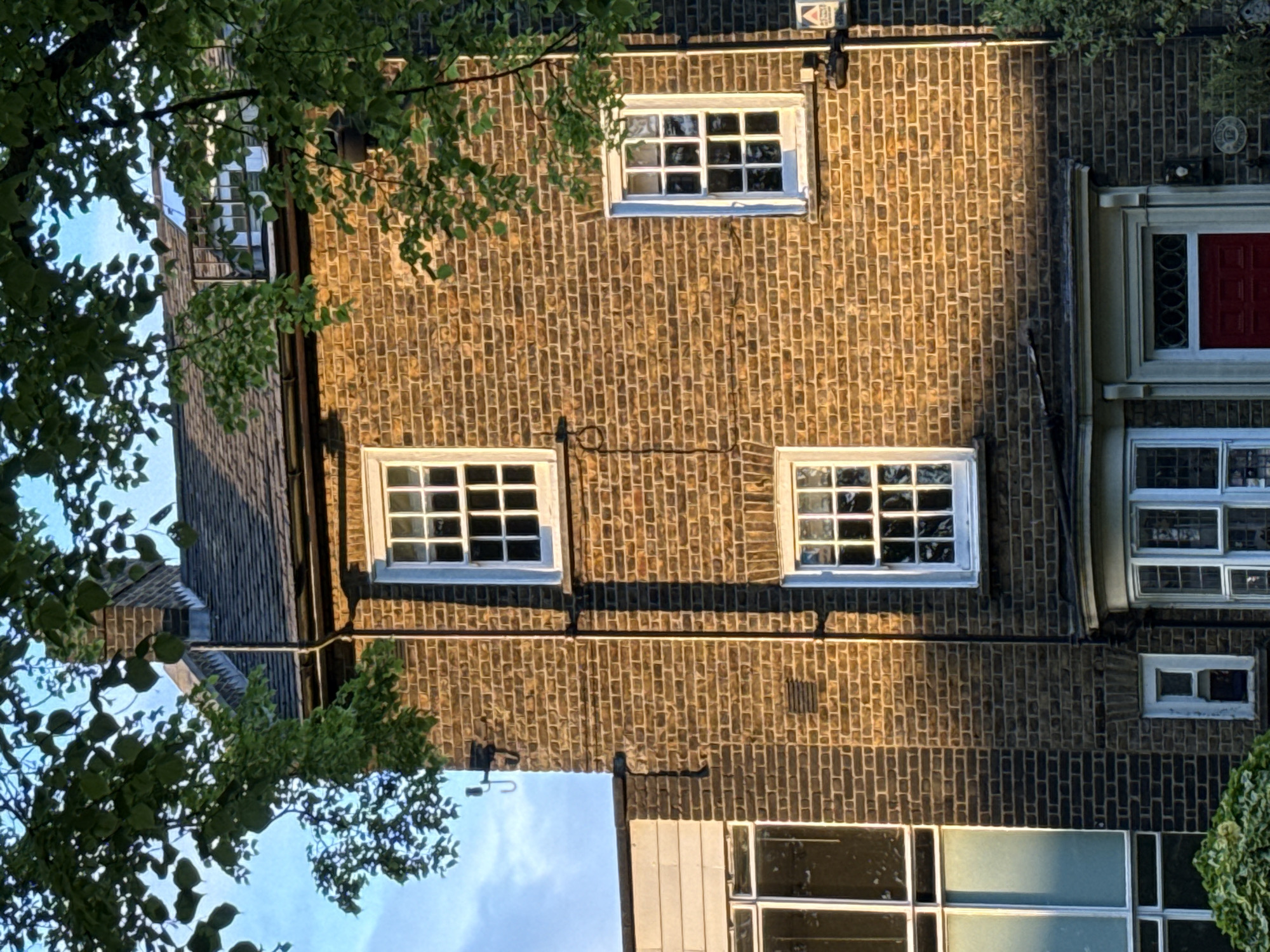 A medium, slightly-tilted, outdoor shot depicts a section of a brick-built building in an urban environment, potentially in London, United Kingdom. The building features multiple windows of varying sizes, most with white frames and divided panes. The brickwork is dark brown with subtle variations in tone, suggesting age and weathering. A black horizontal pipe runs across the brick facade, near the bottom of the shot.
Green foliage, possibly from trees, frames the left and upper edges of the image, partially obscuring the building. The sky is visible in the lower left corner, suggesting a sunny day.
The photograph displays a typical architectural style and design elements found in many residential and commercial buildings in London.