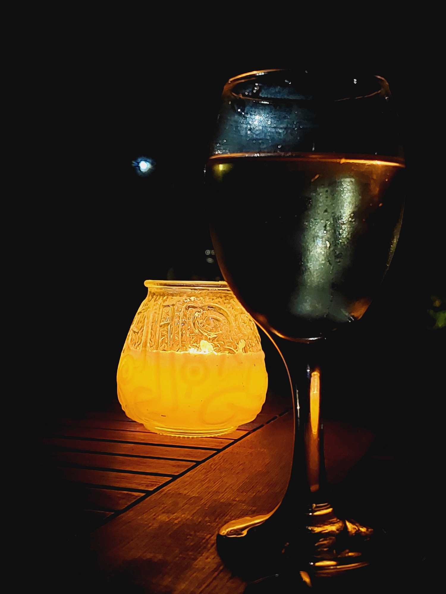 The image depicts a dimly lit outdoor setting, likely a table at a restaurant or cafe in Sozopol, Bulgaria. In the foreground, a wine glass filled with a light-colored liquid is prominently displayed. The glass has condensation on its exterior, suggesting it contains a cold beverage. Behind the wine glass, a candle glows warmly within an ornate, textured glass lantern. The lantern is illuminated by the candlelight, casting a soft yellow glow. The objects are placed on a wooden surface, which appears to be a table. The background is dark, with a faint, out-of-focus light source visible in the upper left corner. The overall mood is serene and intimate, possibly evoking a romantic or relaxing evening atmosphere.