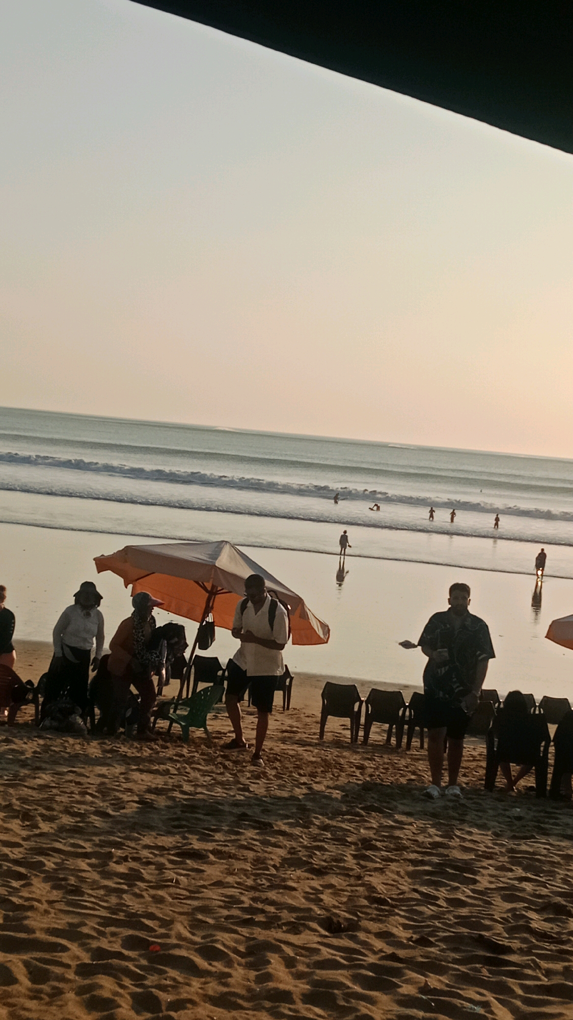 The image depicts a beach scene in Kuta, Indonesia, at dusk. The sun is low in the sky, casting a warm, golden light on the sand and water. The ocean features gentle waves, with several surfers visible in the distance.
In the foreground, a group of people are gathered on the sandy beach. An orange and white umbrella provides shade for some individuals. A man wearing a white shirt and black shorts stands near the umbrella, appearing to be holding something in his hand. To his right, another man in a dark patterned shirt and black shorts stands with his legs apart. Several empty plastic chairs are arranged on the sand, suggesting a waiting area or a place to relax.
Further back, near the water's edge, more people are present. Some appear to be walking or standing, while others are interacting with each other. The overall atmosphere is relaxed and serene, with the setting sun creating a peaceful ambiance.