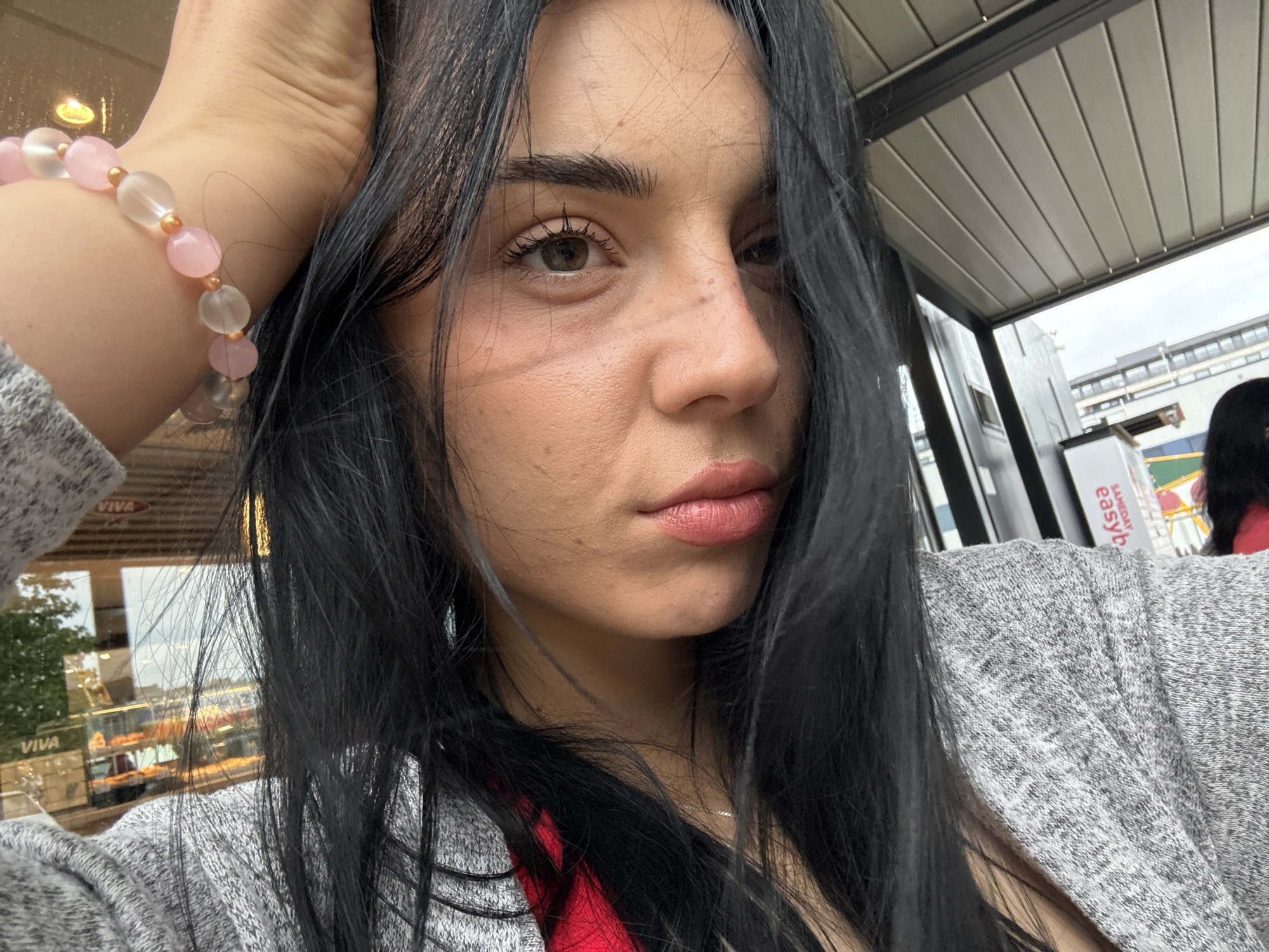A woman with dark hair is posing for a selfie. She's wearing a gray sweater, and a pink and clear beaded bracelet is visible on her wrist. The photo seems to be taken outdoors, near what appears to be a cafe or a shop named "Viva," based on a visible sign. Other signs and building structures are discernible in the background. A sign with the word "easy" in red text is also visible, and a red object. Based on this information and the image context, the location may be in Burgas, Bulgaria.