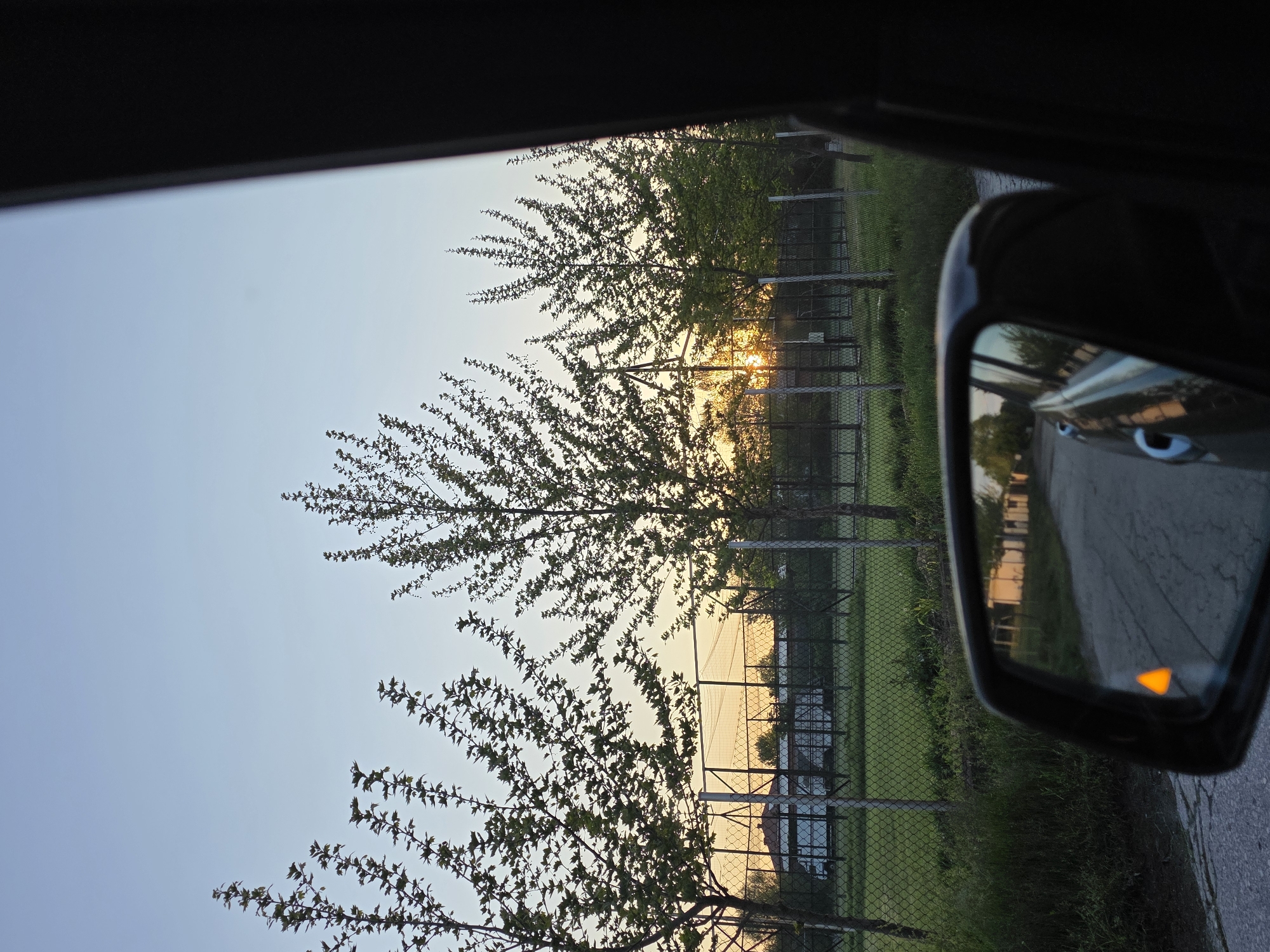 The image depicts a view from inside a car, likely taken while in motion. The focus is on the external environment as seen through the car window and the side mirror.
Through the car window, there are trees with sparse foliage silhouetted against a sky transitioning from blue to a sunset hue. A chain-link fence runs parallel to the trees, with a building visible behind the fence.
In the side mirror, a section of a paved road is reflected along with the side of the car, indicating the car is moving on a road. The turn signal indicator in the mirror is lit.
The location is not discernible from the image, and there is no verifiable information to confirm the location is in Yagodovo, Bulgaria. There is no indication of people in the photo.