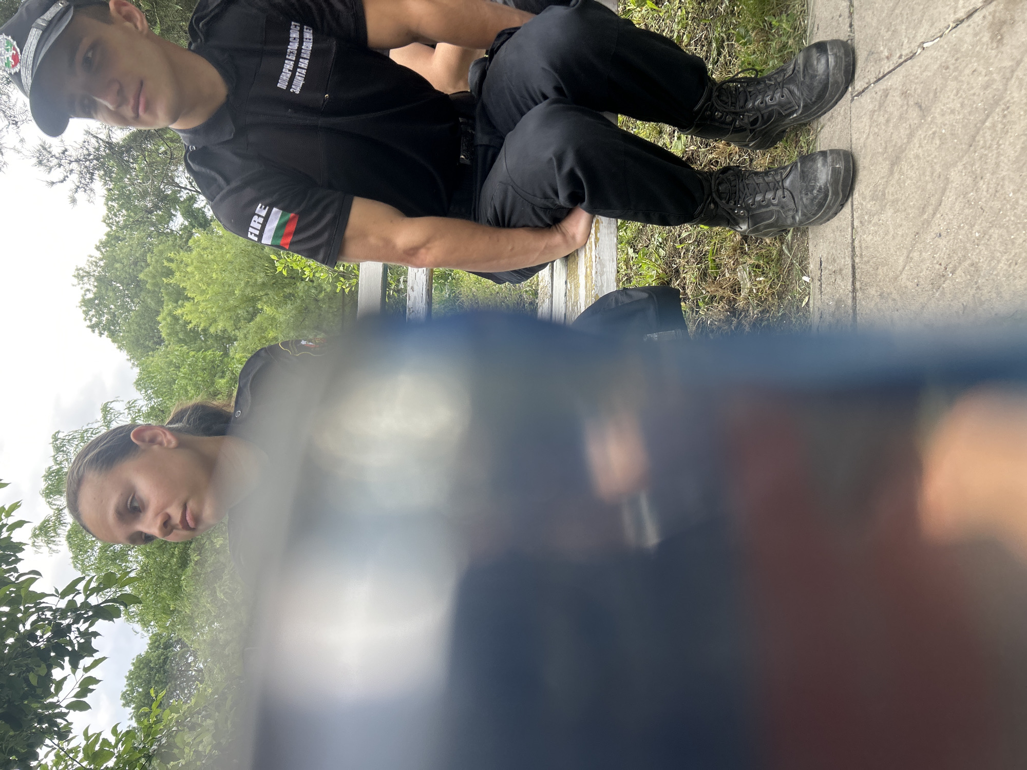 The image shows two people, one male and one female, likely sitting on a bench outdoors. The male is wearing a black uniform with patches that appear to indicate Bulgarian "FIRE" services, including a Bulgarian flag. He is also wearing a hat. He is sitting in a relaxed posture. The female is partially obscured and positioned lower in the frame. The background includes greenery and trees. Given the uniform details, the location is plausibly in Sofia, Bulgaria. The scene appears casual, suggesting a break or moment of rest.