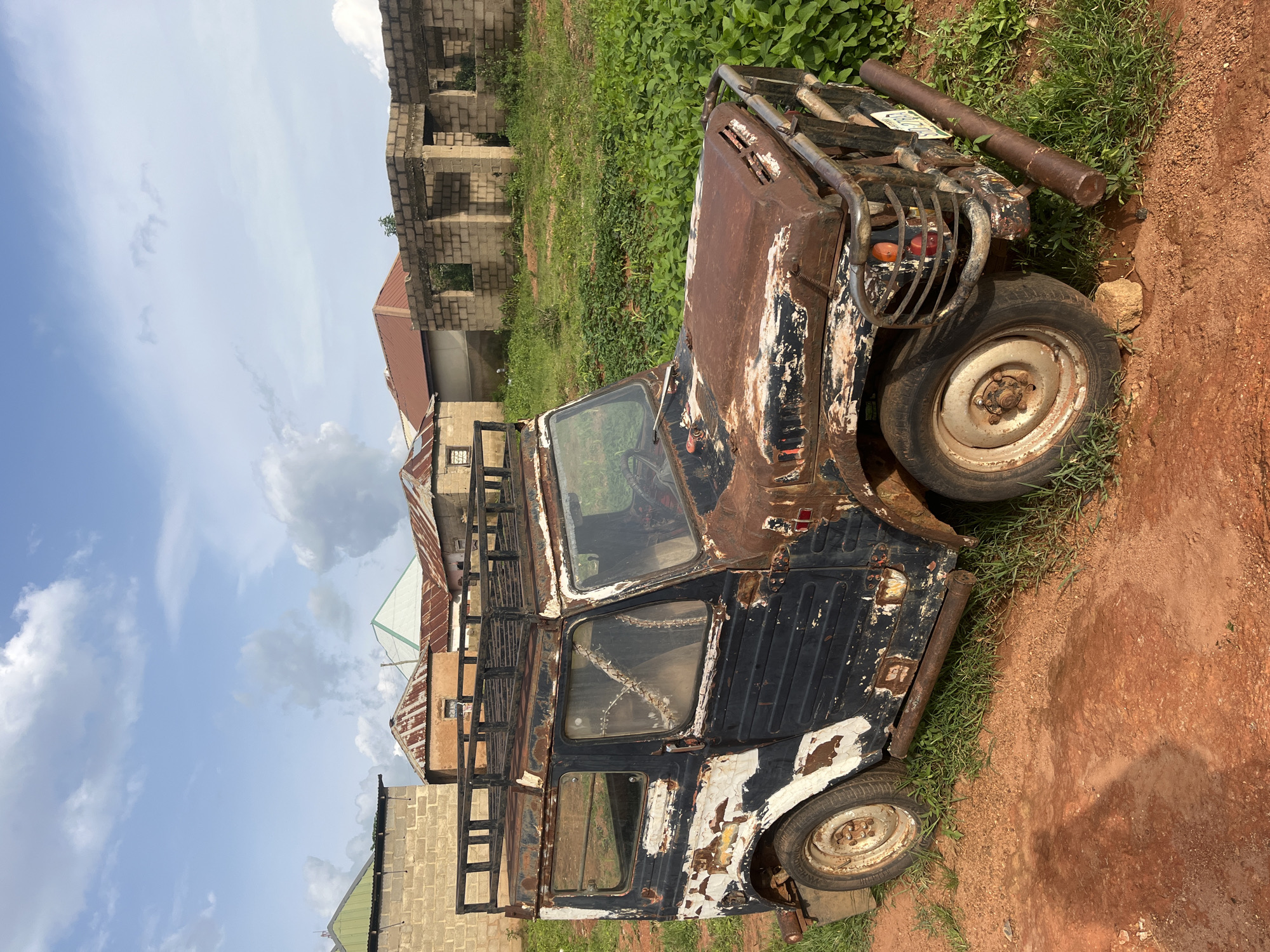 A rusted, dark blue and white jeep with a roof rack sits on a dirt road in Unguwar Saya, Nigeria. The jeep is in disrepair, with peeling paint and visible rust. In the background, there are several unfinished brick buildings and houses with corrugated metal roofs. Green vegetation surrounds the area. The sky is partly cloudy with white cumulus clouds.