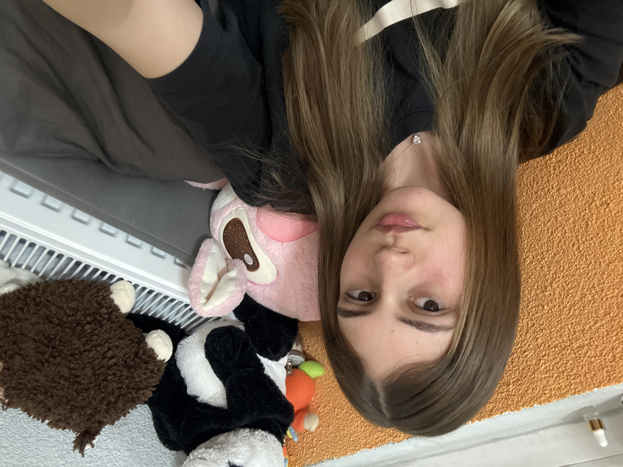 A young woman with long brown hair lies on her side on an orange textured surface, possibly a towel or blanket. She is wearing a black shirt with a white stripe on the shoulder and a delicate necklace with a heart-shaped pendant. Several stuffed animals are clustered near her: a brown hedgehog, a black and white cow, and a pink bunny. A white radiator with a gray shelf above it is visible in the background. A small, clear bottle with a white and gold lid lies near the edge of the orange surface. The location is not evident in this image.