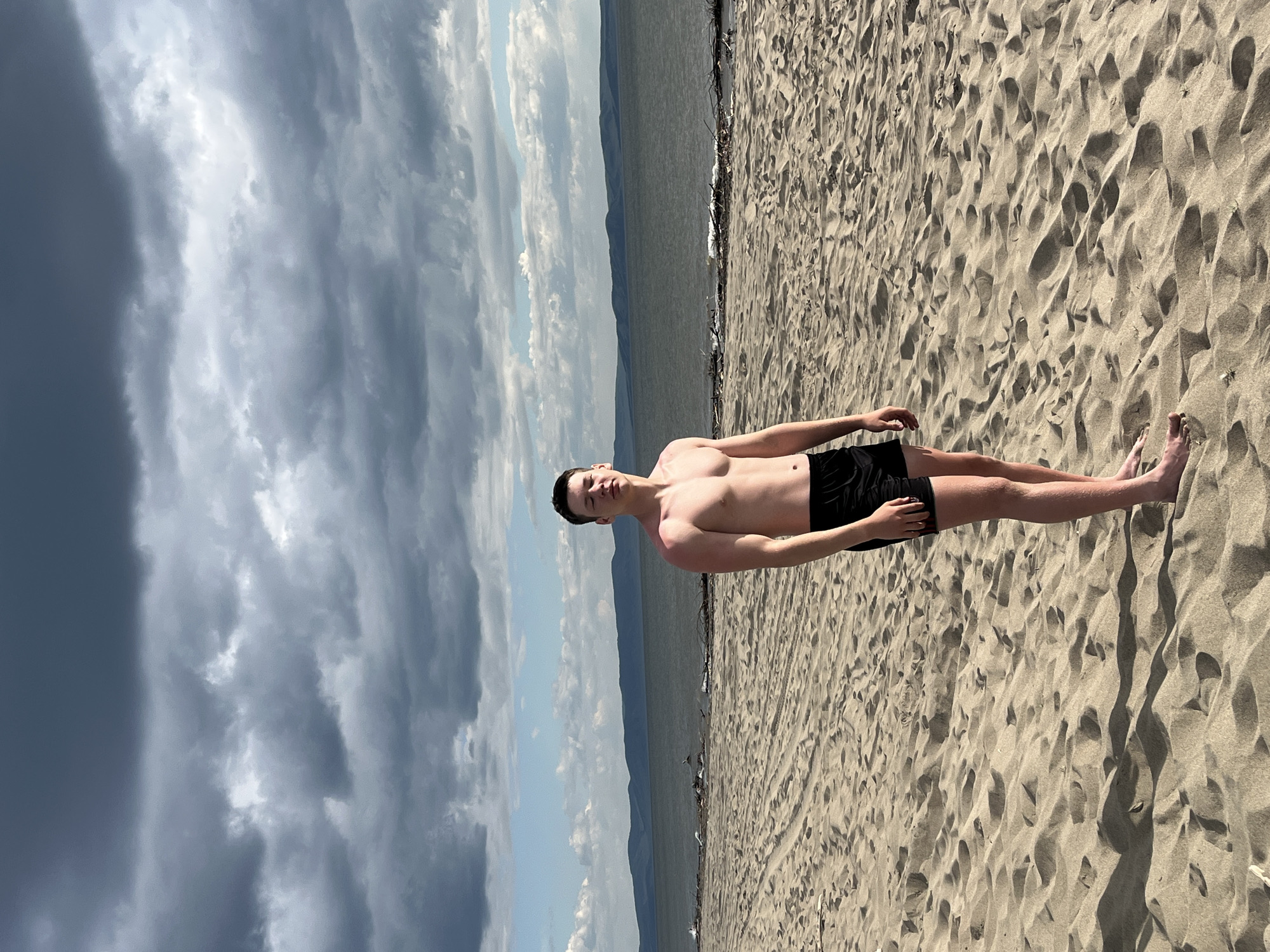 The media file shows a young man, shirtless and wearing black swim trunks, standing on a sandy beach. He is facing the camera with his arms relaxed at his sides. The beach is covered in fine, light-colored sand with distinct ripples and textures. In the background, a body of water stretches out, meeting a distant mountain range under a dramatic sky. The sky is filled with a mix of dark, brooding storm clouds on the left and brighter, lighter clouds with patches of blue sky on the right, suggesting a dynamic weather front. A few birds are scattered on the beach near the water's edge. The scene appears to be set during the daytime, given the lighting conditions and shadows cast on the sand. The overall mood is casual and serene, despite the imposing clouds. There is no visible text in the image. The location context is Krasnoturansk, Russia.
