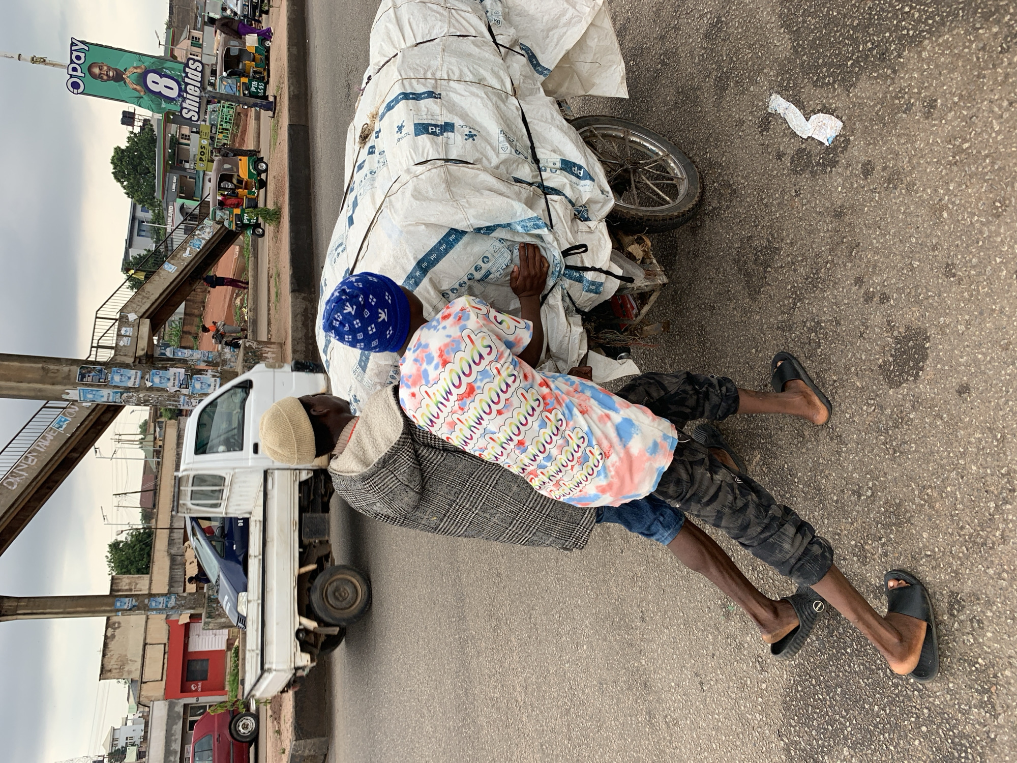 This is a photograph taken in Gura Top, Nigeria, likely during the daytime. The scene is a roadside with a busy atmosphere.
In the foreground, two individuals are lying on the asphalt next to a large, white, tarp-covered object, possibly a motorcycle or cart laden with goods. One person is wearing a patterned shirt with the word "Suchwoods" repeated in rainbow colors, and a grey checkered jacket. They also have a blue patterned hat. The other person is partially obscured but appears to be wearing camouflage-patterned trousers and black slides. Their body language suggests they are resting or possibly asleep.
Behind them, a white truck is parked, and further back, a pedestrian bridge can be seen. There are also several three-wheeled vehicles, commonly known as Keke Napep, on the road. A large billboard advertising "OPay" and "8 Shields" is prominently displayed in the background, along with other smaller signs. The ground is asphalt, with some patches of wetness and scattered debris. The sky is overcast, suggesting cloudy weather. The overall impression is one of a typical urban street scene in Nigeria, with commercial activity and everyday life unfolding.