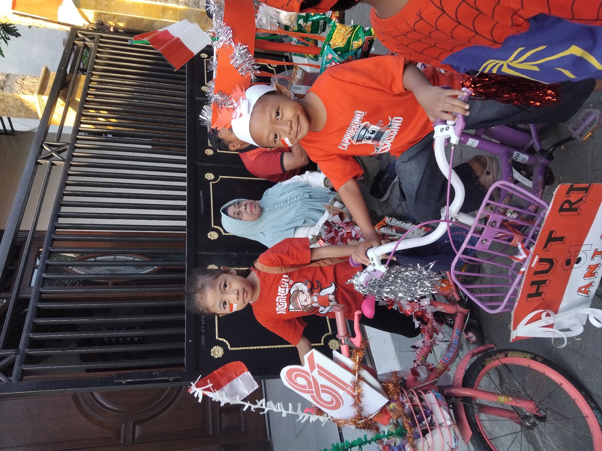 In Kepuh Kiriman, Indonesia, a group of children and adults are gathered outdoors, possibly for a local celebration or parade. Two children are prominently featured, each with Indonesian flags painted on their cheeks. The child on the left, a girl, is wearing a red t-shirt with a cartoon graphic and is standing next to a pink bicycle adorned with Indonesian flags and decorations. The child on the right, a boy, is wearing an orange t-shirt with a coffee-related graphic and a white paper crown, and is sitting on a purple bicycle decorated with red tinsel. Behind them, an adult woman in a light blue hijab and glasses, and an adult man in a red shirt are visible. The background consists of a house with a black metal fence and a large wooden door. The overall scene suggests a festive and patriotic atmosphere.