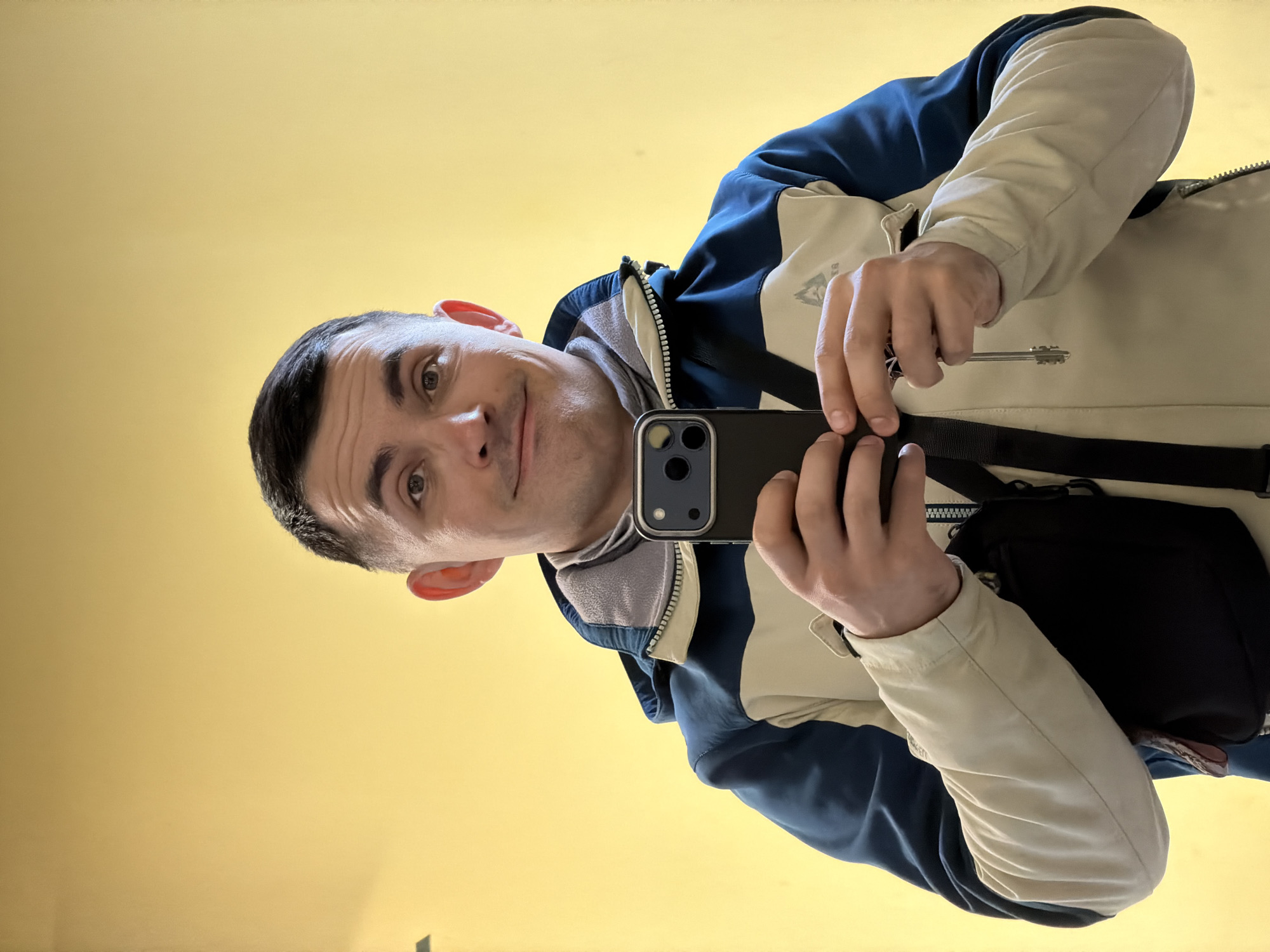 A man is taking a selfie in what appears to be an indoor setting with a yellow background. He is holding a black smartphone with visible camera lenses. The man is wearing a jacket with a white top section and navy blue lower section. A gray hooded sweatshirt is visible underneath. He has short dark hair and is looking at the camera with a slight smile. A key is partially visible in his left hand. A black bag strap is across his body. The location in the city of Izhevsk, Russia, cannot be determined from the image.