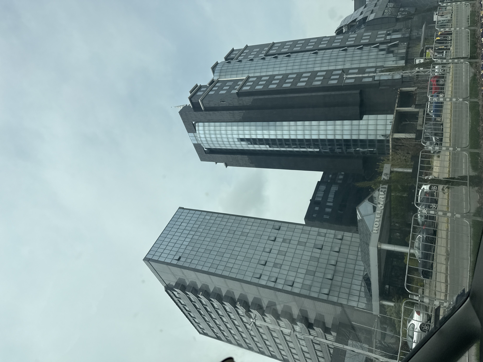 The image depicts two modern glass and concrete buildings against a cloudy sky. A sign reading "HOTEL MART" is visible, indicating the hotel's location in Plovdiv, Bulgaria. Cars are parked along the street beside the buildings. A reflective metal fence is partially visible. The photo appears to be taken from inside a vehicle.