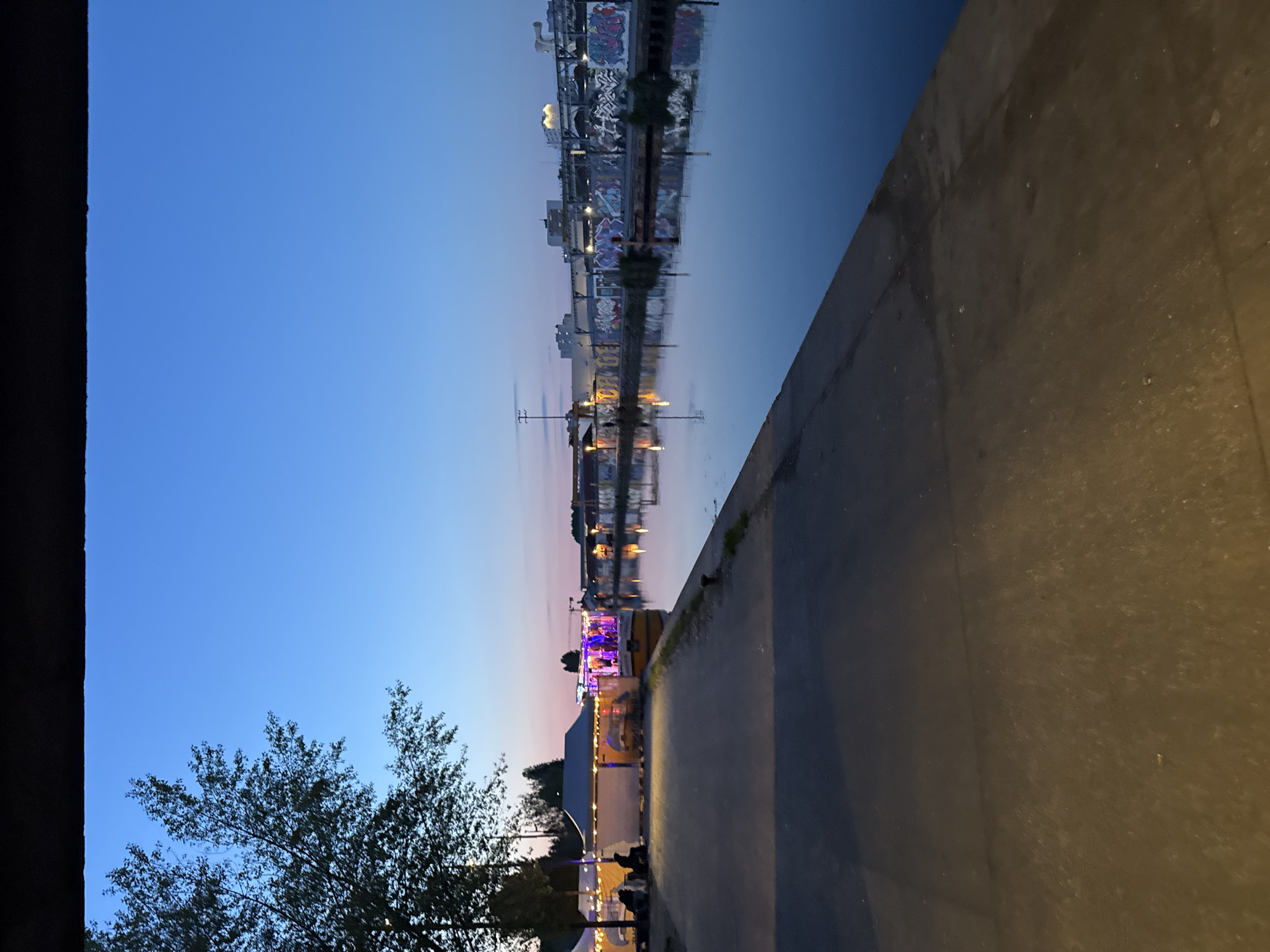 The image shows a view of the Canal Saint-Martin in Paris, France, at dusk. The sky transitions from a deep blue to a soft orange-pink near the horizon. On the right side of the frame is a concrete embankment bordering the canal. The water surface reflects the city lights and the buildings across the canal. A tree with bare branches is visible in the lower-left corner. Along the canal bank, there are tents, boats, and buildings illuminated with light.