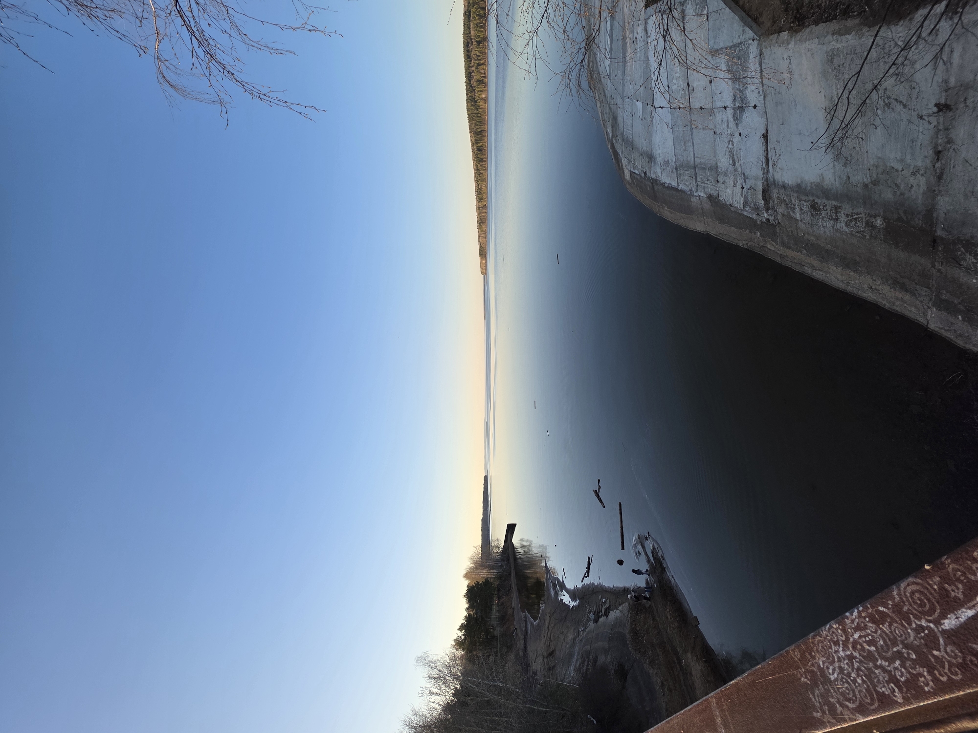 The image shows a view of the Kama Reservoir in Chaikovsky, Russia. A curved concrete dam structure is visible on the right. Calm water fills much of the scene, reflecting a pale sky. A thin strip of forest is seen on the distant shore. In the lower left, there's an area of shoreline with logs and vegetation. A metal railing with decorative elements is in the foreground. The sky is predominantly blue with a gradient toward the horizon. Bare branches from trees are visible in the upper left and right corners.