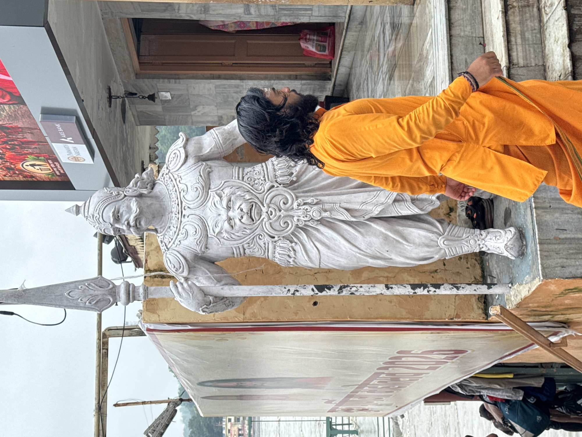 A man in an orange garment walks down steps near a large white statue of a warrior in Rishikesh, India. The statue is adorned with ornate armor and a lion's face on its chest. The man has long, dark, wavy hair and a beard, and is wearing a traditional Indian outfit. Behind the statue, a large banner with images and text in Hindi is visible. The background shows a building with signs for "Axis Bank" and "Parmarth Niketan." In the distance, a river can be seen. The scene appears to be at an outdoor location, possibly near a temple or ashram.