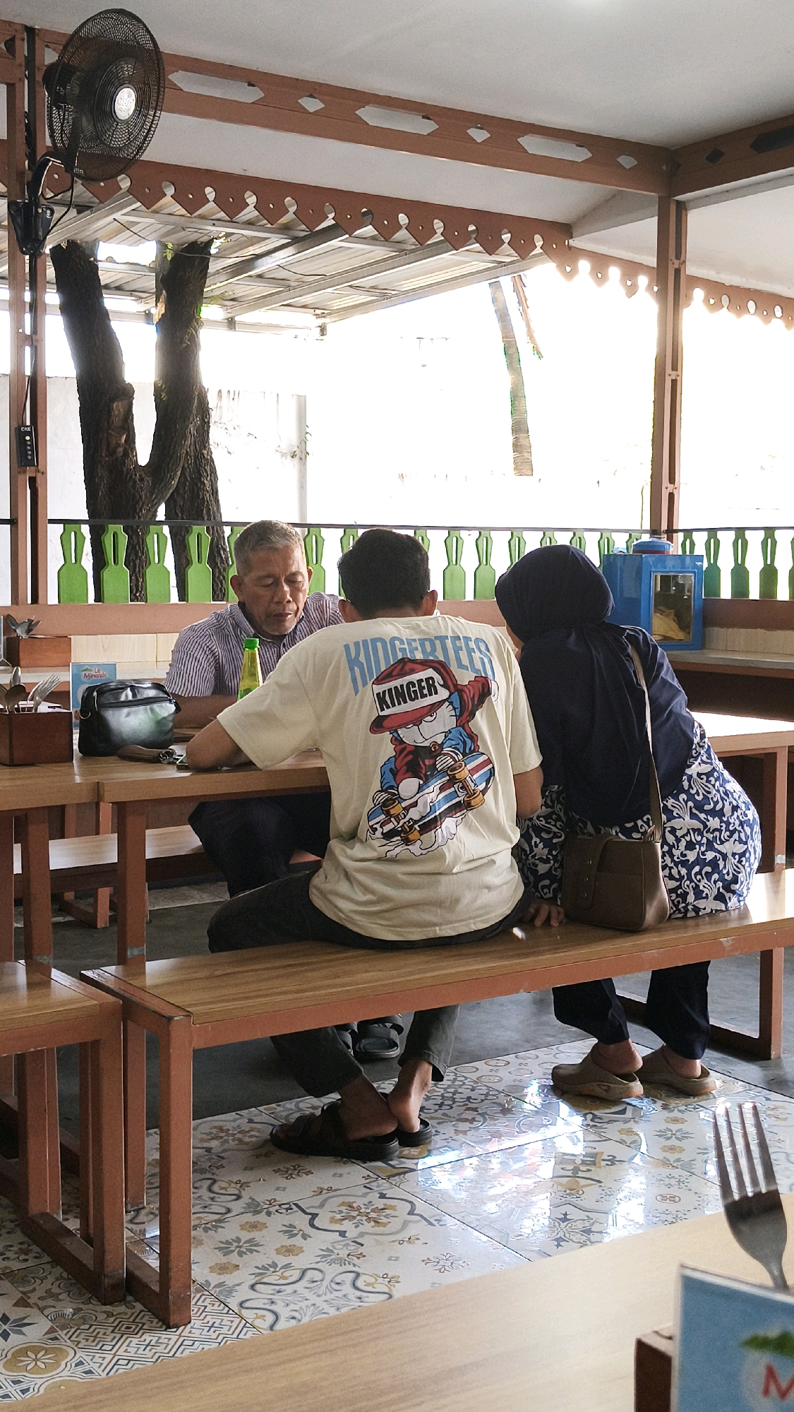 Three individuals are seated at a wooden table in an open-air eatery, possibly a cafe or restaurant, in Bekasi, Indonesia. An older man with short grey hair is seated on the left, wearing a striped collared shirt. He is looking down at the table, and a green bottle is placed in front of him. To his right is a younger man wearing a cream-colored t-shirt with a graphic print of a cartoon character on a skateboard and the text "KINGERTEES KINGER" on the back. He is also looking down. On the right side of the table, a woman wearing a dark hijab and a patterned blue and white skirt is seated. She has a brown shoulder bag on her right side. The flooring is tiled with a decorative pattern. In the background, there are green decorative elements on a wall, a ceiling fan is mounted overhead, and trees are visible outside. The overall lighting suggests it is daytime.