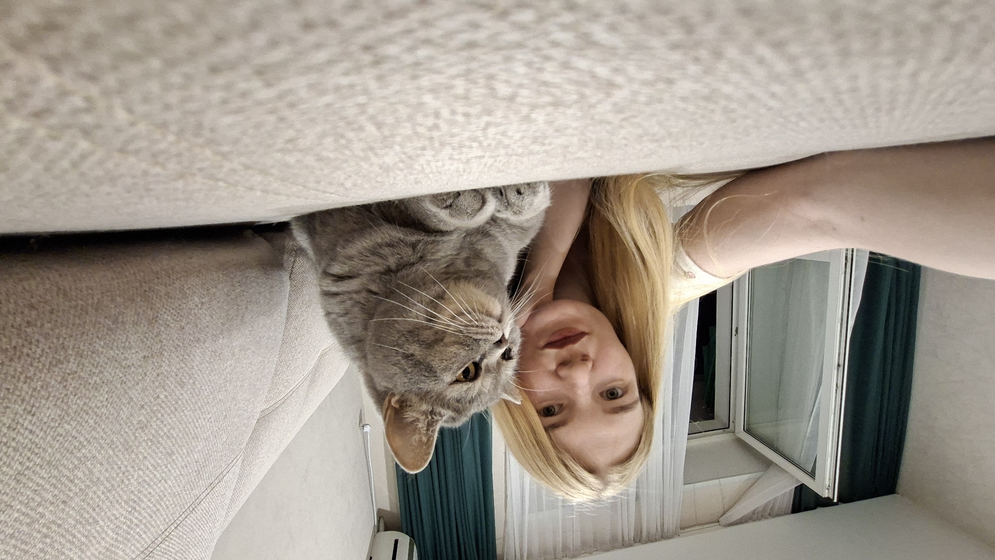 An adult female and a domestic cat are situated on a light-colored, textured fabric sofa in an interior residential setting in Chaikovsky, Russia. The female, with long blonde hair, light complexion, and dark lipstick, lies on her stomach, propped on her right forearm with her left hand supporting her chin. She is wearing a light-colored top and looks towards the camera, which is positioned low on the sofa. Adjacent to her, a grey British Shorthair cat with amber eyes is lying with its front paws tucked, facing right and gazing off-camera. In the background, a white-framed window or balcony door is partially open, flanked by dark teal curtains on the left and white sheer curtains on the right. A light-colored wall with a white wall-mounted air conditioning unit on the upper right is visible behind the sofa.