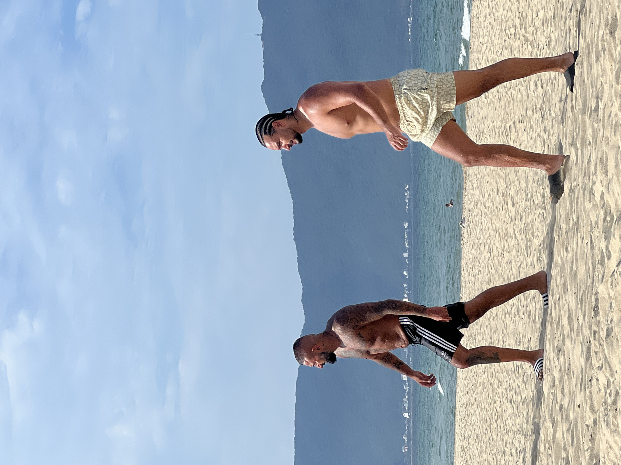 Two shirtless men walk along a sandy beach in Da Nang, Vietnam. The man on the left has a shaved head, a beard, and numerous tattoos on his torso and arms. He is wearing black shorts with white stripes and white slides. The man on the right has his hair in braids, a beard, and is wearing light-colored patterned shorts and dark slides. In the background, the ocean meets a shoreline with buildings, and beyond that, hazy mountains rise under a pale blue sky with scattered clouds.