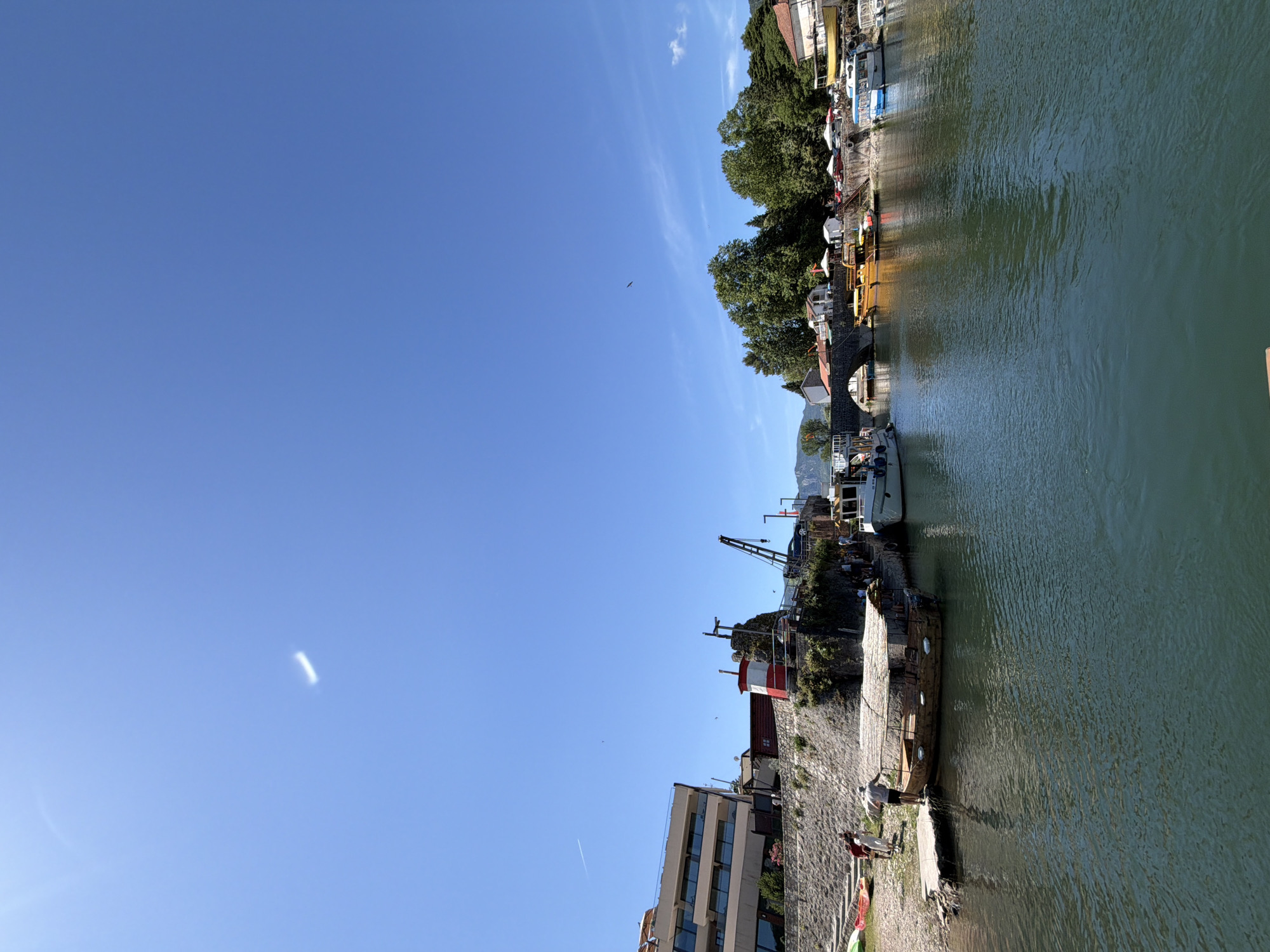 The image depicts a daytime scene along a river or canal in Boljevići, Montenegro, under a clear blue sky. The sky features a faint white contrail in the upper left quadrant and a small, luminous, rectangular artifact near the top center, possibly a lens flare. A large bird is visible in flight in the upper right quadrant.
The left bank features a prominent stone retaining wall. Above this wall, a multi-story building with balconies is visible on the far left, appearing to be a modern hotel or residential structure. Closer to the water, a red and white striped cylindrical structure, resembling a small lighthouse or marker, stands on the wall, accompanied by a dark-colored crane structure further right. Below the wall, a small pebbled beach area is visible with two kayaks (red and yellow) resting on the shore. Several individuals are present on this beach. One person, wearing a grey shirt and white hat, stands on a small concrete pier, appearing to interact with a wooden boat. Another person in a red shirt is nearby. A rustic wooden boat with a covered roof is moored alongside the concrete pier. A modern white boat is partially visible moored further along the left bank.
The right bank features a stone arch bridge spanning the water. Structures with orange-tiled roofs are visible above the bridge. The bank is heavily vegetated with numerous large green trees. Beyond the trees, several buildings with red roofs are discernible, along with small white tents or umbrellas, and parked vehicles. Several boats are moored along the right bank, including a bright yellow boat and a blue boat.
The water itself is a dark green-blue, displaying ripples across its surface. Distant mountains are visible on the horizon, framing the scene.