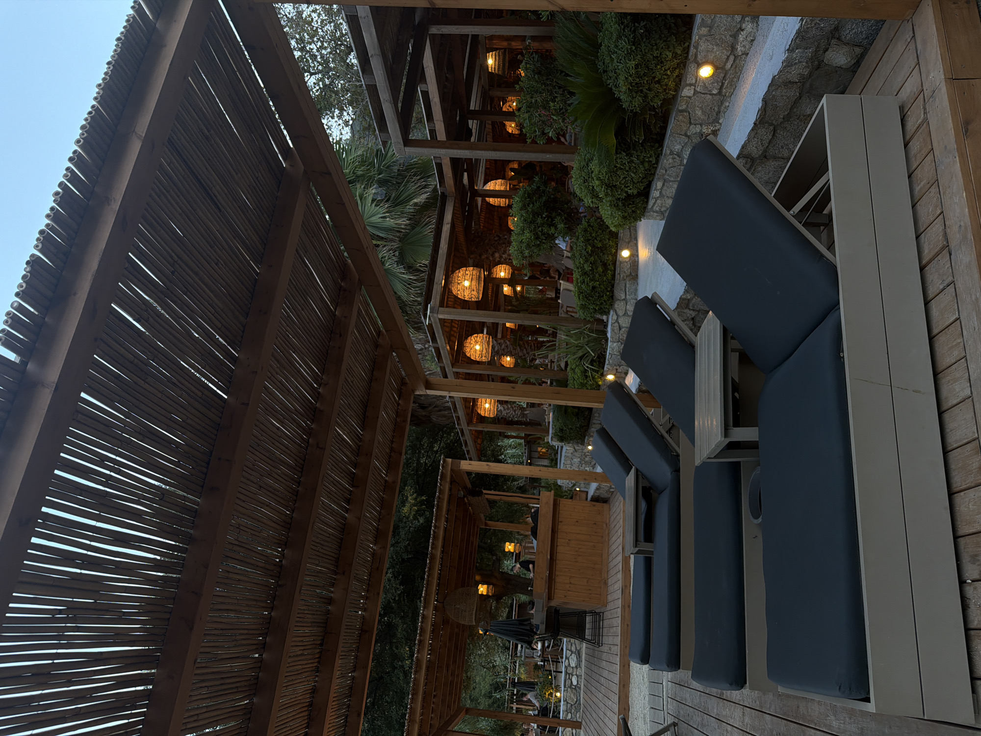 The image depicts an outdoor seating area at a beach resort in Sithonia, Greece. In the foreground, there are several dark-colored lounge chairs arranged on a wooden deck. The chairs are partially covered by a bamboo-thatched roof supported by wooden beams. Beyond the lounge chairs, there is a bar area with stools and patrons seated at tables. The background features lush greenery, palm trees, and more covered seating areas with woven lamps. The scene appears to be set in the early evening or late afternoon, with ambient lighting from the lamps illuminating the area.