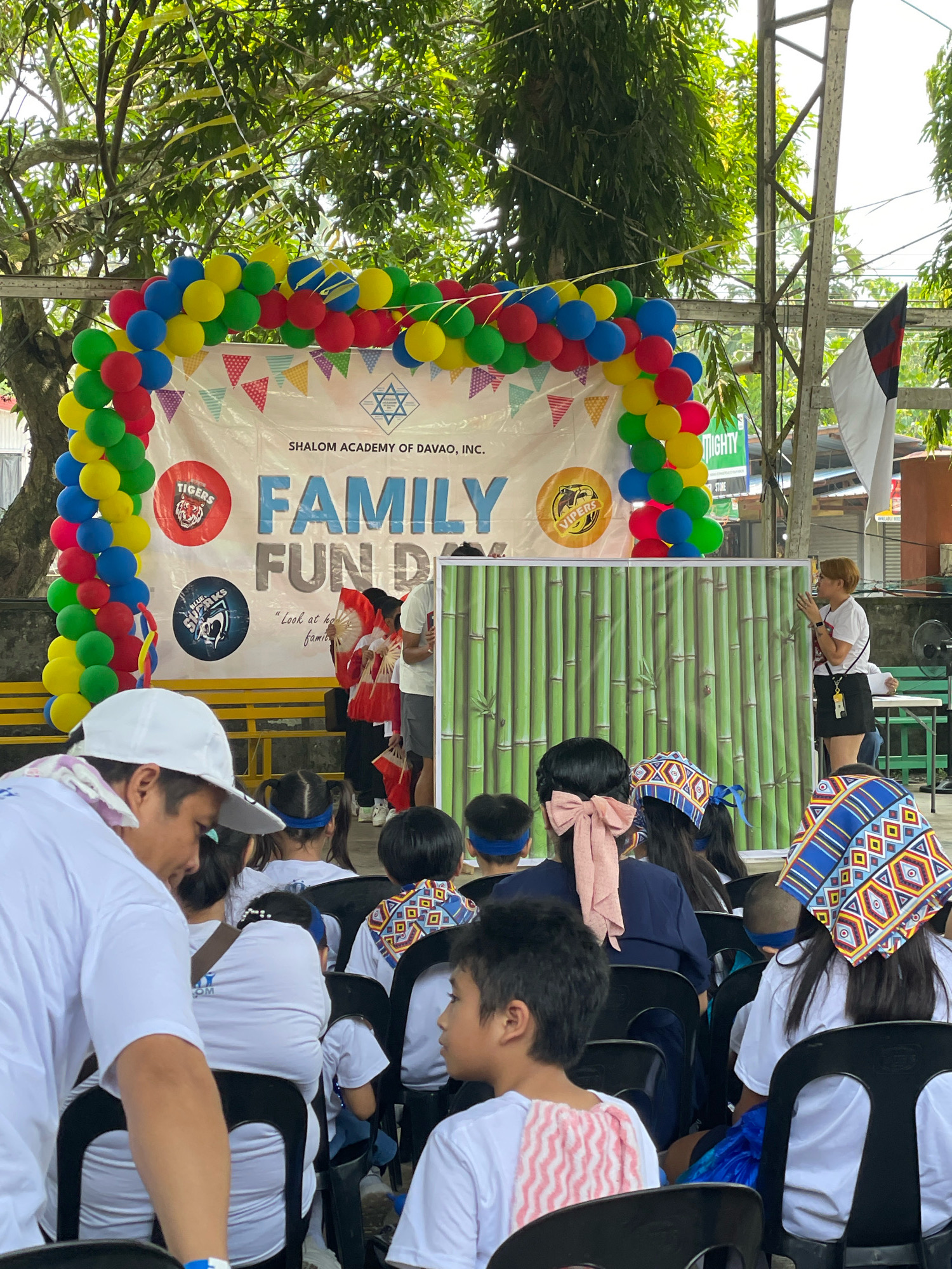 This is a medium shot photograph taken outdoors during daytime, capturing a family fun day event organized by Shalom Academy of Davao, Inc. The event appears to be held in an open area, possibly within the school grounds or a community park in Davao City, Philippines, given the signage.
In the background, a large banner displays "FAMILY FUN DAY" in bold blue lettering, with "SHALOM ACADEMY OF DAVAO, INC." above it. The banner is partially framed by a colorful arch of balloons in red, yellow, green, and blue. Two circular logos are visible on the banner: one featuring a tiger and the other with "CLUB VIPERS" and a snake. A decorative bamboo-print backdrop is set up on a stage.
In the midground, several individuals are present. A woman with short, light brown hair and wearing a white t-shirt and black skirt is addressing a group. She is holding a small object in her right hand and gesturing with her left. On the stage, two individuals in traditional red and white attire with fans are performing or posing.
The foreground is occupied by an audience seated on black chairs, mostly children. Many of the children are wearing white t-shirts and some have headbands or headscarves. One child in the far right foreground is wearing a blue, yellow, and white patterned headscarf. Another child in the center foreground has a large pink bow in their hair. A person wearing a white cap and a white shirt, partially obscuring the view of the children, is in the left foreground. The environment suggests a warm climate, with lush green trees visible above and behind the banner. A flag, predominantly white with black and red elements, is attached to a metal pole on the right side of the frame.