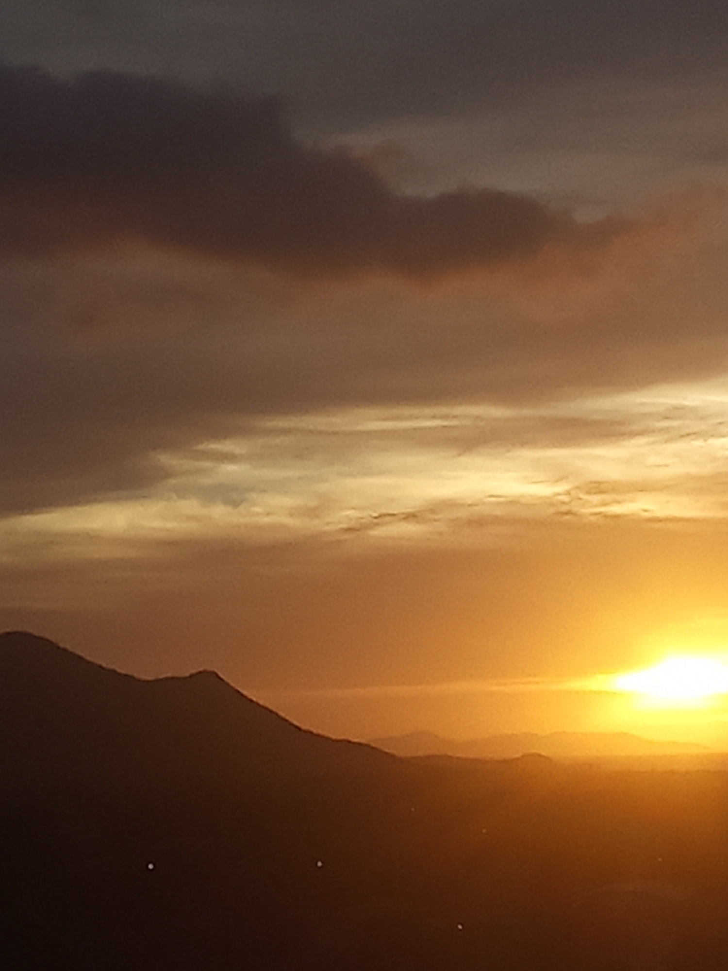 The image displays a sunset or sunrise over a mountainous landscape, specifically located in Juan Griego, Venezuela. The scene is dominated by a bright, intense yellow-white sun positioned in the lower right quadrant, casting strong golden and orange light across the lower sky. The sky transitions from dark grey and brown clouds in the upper left to lighter, striated clouds infused with golden and orange tones across the mid-section. A range of mountains is silhouetted in dark brown to black across the lower third of the frame, showing distinct peaks and undulating contours. Fainter, receding mountain layers are visible within the atmospheric haze towards the horizon. Two small, bright white circular points of light are visible within the dark silhouette of the foreground mountain range. There are no individuals, explicit human actions, or distinct man-made objects discernible within the frame, apart from the two potential distant lights.