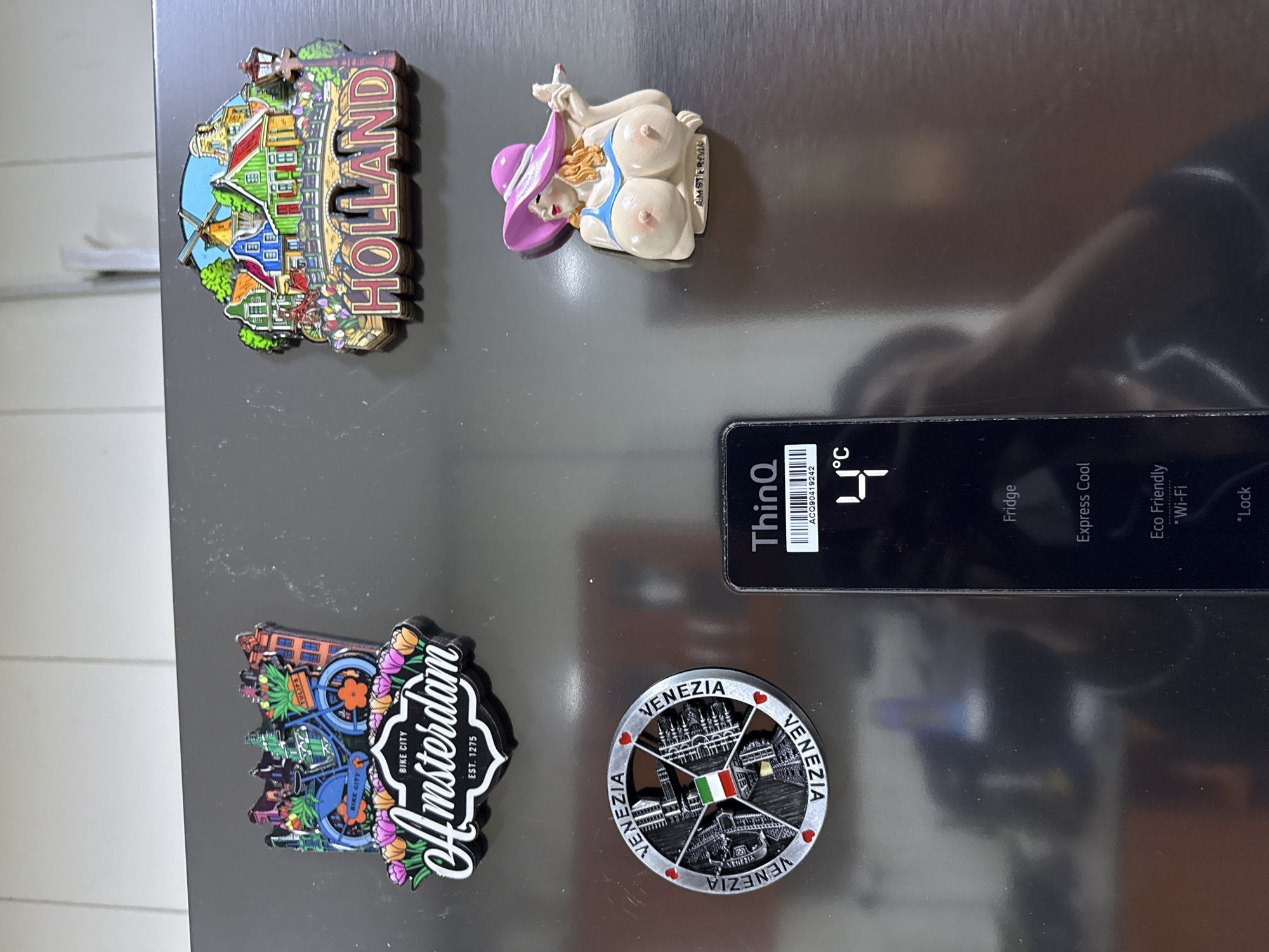 A close-up, vertical photograph captures the upper-mid section of a dark metallic, likely stainless steel, refrigerator door, situated within an indoor setting in Alaguntan, Nigeria. The refrigerator surface is adorned with four distinct magnets and features an integrated digital control panel.
In the upper-left quadrant, a rectangular magnet depicts a blue bicycle laden with flowers, set against a backdrop of colorful city buildings, with the text "BIKE CITY Amsterdam EST. 1275" below. Adjacent to it on the upper right, another rectangular magnet portrays a quintessential Dutch landscape featuring colorful houses, a windmill, a bridge, and tulips, with "HOLLAND" prominently displayed at the bottom.
Below the Amsterdam magnet, a circular silver-colored magnet features a stylized depiction of Venice, including architectural elements and the Italian flag at its center, encircled by repetitive "VENEZIA" text and small red hearts. To the right of the Venezia magnet, a three-dimensional figurative magnet portrays the upper torso of a woman with fair skin, wearing a pink wide-brimmed hat and a light blue top, exposing prominent breasts with defined nipples. She holds a white cylindrical object, consistent with a cigarette, towards her mouth area with her left hand.
A vertical black digital control panel, branded "ThinQ", is positioned on the lower-right half of the refrigerator door. The screen displays "4°C" as the current temperature and lists menu options including "Fridge," "Express Cool," "Eco Friendly," "Wi-Fi," and "*Lock." A barcode labeled "ACQ90419242" is visible at the top of the display. The reflective surface of the refrigerator exhibits blurry reflections of the immediate indoor environment and a partial, indistinct reflection consistent with an individual holding a camera. The background above the refrigerator consists of a light-colored wall with vertical seam lines.
