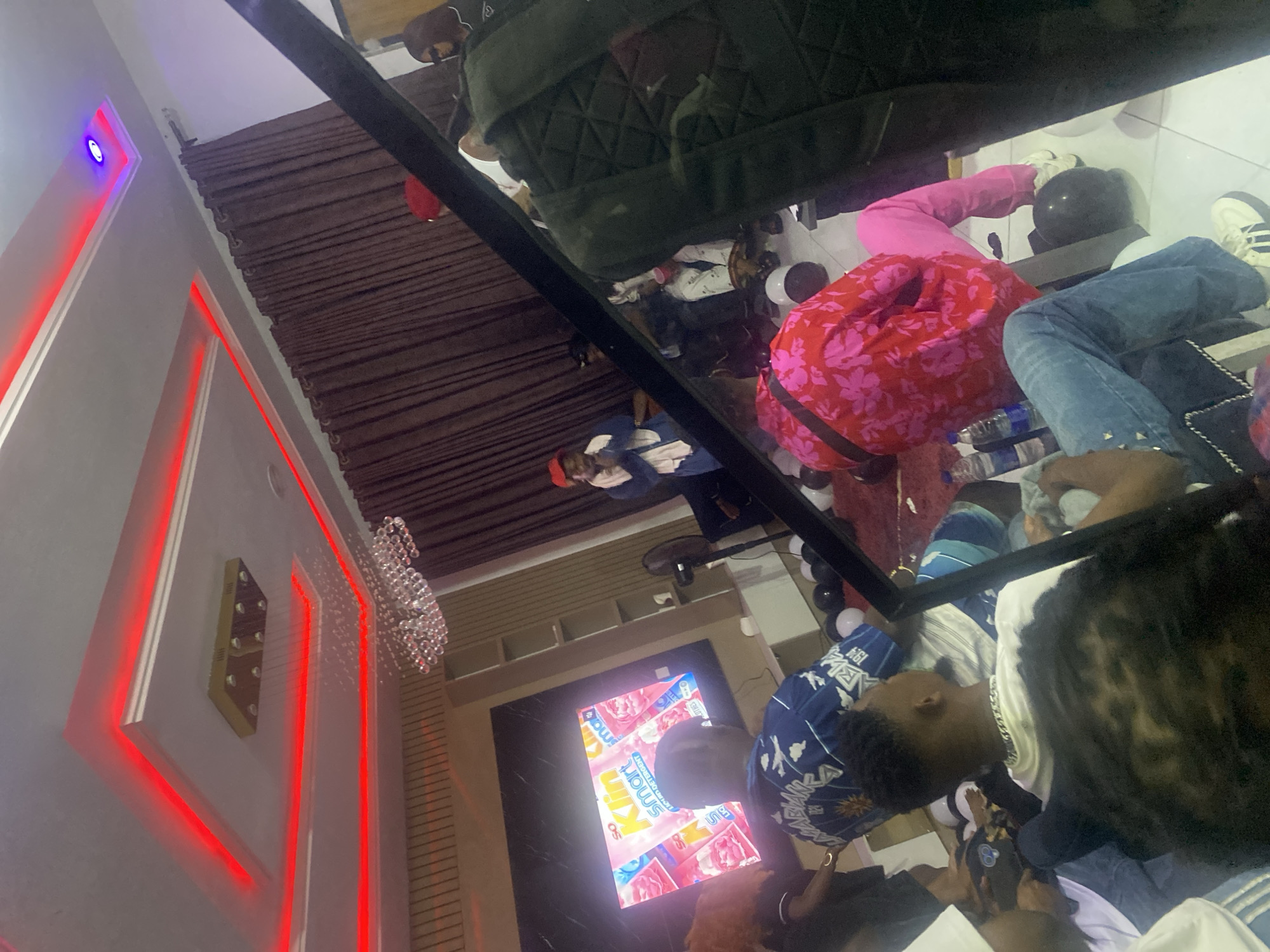 The image depicts an indoor gathering in a modern room located in Lekki, Nigeria. The ceiling is white with a tiered false ceiling design, featuring prominent red LED strip lighting and a central crystal chandelier, along with a smaller square gold light fixture and a single purple LED spotlight. The rear wall is covered by dark brown, floor-length curtains. The left wall exhibits horizontal wooden paneling. The floor consists of white tiles with dark grout lines.
A large flat-screen television is mounted on the left wall, displaying an advertisement for "So Klin Smart Liquid Detergent." A tall, dark standing fan is positioned to the right of the TV. Scattered across the white tiled floor are numerous black and white balloons.
At least ten individuals are present in the room. A male figure stands centrally towards the rear, wearing a red cap and a light-colored wrap over a dark shirt, appearing to address the group. To his immediate right, partially obscured, is an individual in a dark hoodie and sunglasses. In the midground, a person is seated on the floor, facing away from the camera, dressed in a red and pink floral shirt paired with pink pants and white sneakers.
In the foreground, a person with dark, textured hair is partially visible. Next to them, another individual, seen from behind, wears a white t-shirt, blue jeans, a silver chain necklace, and white sneakers, holding a white object. Further left, a person is visible from behind, wearing a blue and white patterned short-sleeved top. Other individuals are partially visible or obscured throughout the scene, seated on the floor or standing. Bottled water is visible on the floor near some attendees. A dark, rectangular, structural element partially obscures the lower right foreground.