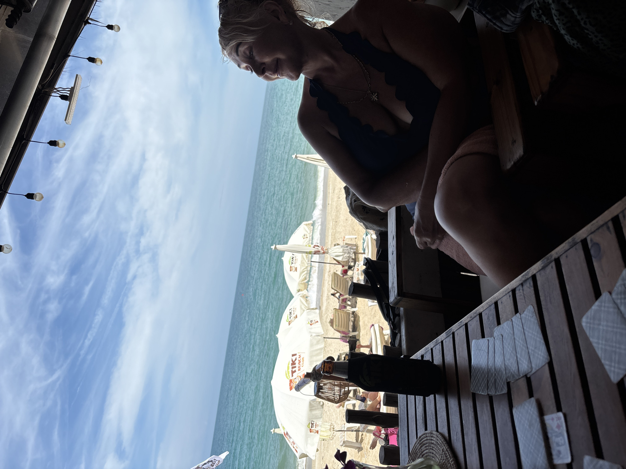 The image shows a woman in a navy swimsuit seated at a wooden table with playing cards, situated at a beachside location in Baan Hua Thanon, Thailand. The scene overlooks a beach with multiple white sun umbrellas marked with 'TIKI BEACH CLUB', lounge chairs, and people relaxing on the sand. The ocean stretches into the background, with a cloudy blue sky above. A string of lights hangs overhead.