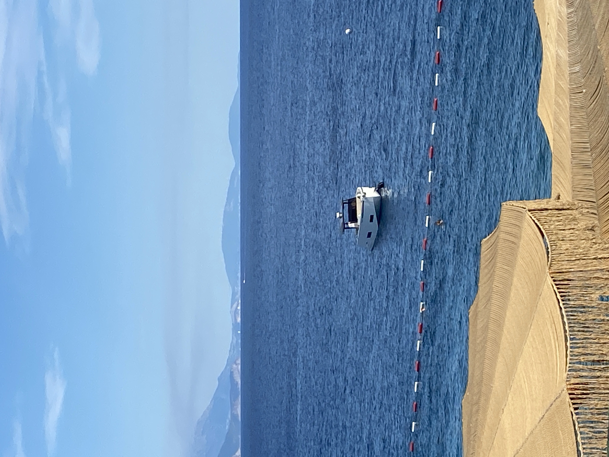 A small, white motorboat with a dark hardtop is moored in the calm, deep blue waters of the Adriatic Sea. The boat is positioned centrally in the frame, with a white buoys in the water to its right. A line of red and white buoys, marking a swimming area, stretches across the foreground of the sea. In the far distance, hazy, blue-toned mountains form the horizon, beneath a pale blue sky with scattered white clouds. In the foreground, the edge of a thatched-roof beach umbrella or cabana with a fringed edge is visible, its textured surface made of woven straw-like material. The scene appears to be a tranquil day at the beach near Prijevor, Montenegro.