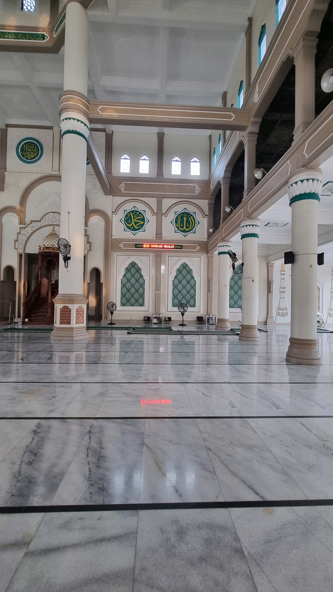 The image captures the interior of the Baiturrahman Grand Mosque in Lhokseumawe, Indonesia, on a well-lit day. The scene is dominated by the mosque's architecture, characterized by its spacious prayer hall with polished marble flooring and tall, fluted pillars supporting an intricate ceiling. The pillars are adorned with green and white decorative bands.
The prayer hall features a mihrab and mimbar on the far wall, with Islamic calligraphy and decorative archways above. Several framed Islamic calligraphy artworks are prominently displayed on the walls. Two large, ornate windows with green patterned screens are visible, adding to the visual appeal.
In the foreground, the marble floor reflects the ambient light and the architectural elements of the mosque. There are several black standing fans placed strategically across the hall. Prayer mats are visible in various locations. A digital display board with text in Malay, likely indicating prayer times, is mounted on the wall below some of the calligraphic artworks.
The overall impression is one of serenity and grandeur, typical of a significant religious building. The image appears to be taken from a low angle, emphasizing the scale and height of the mosque's interior.