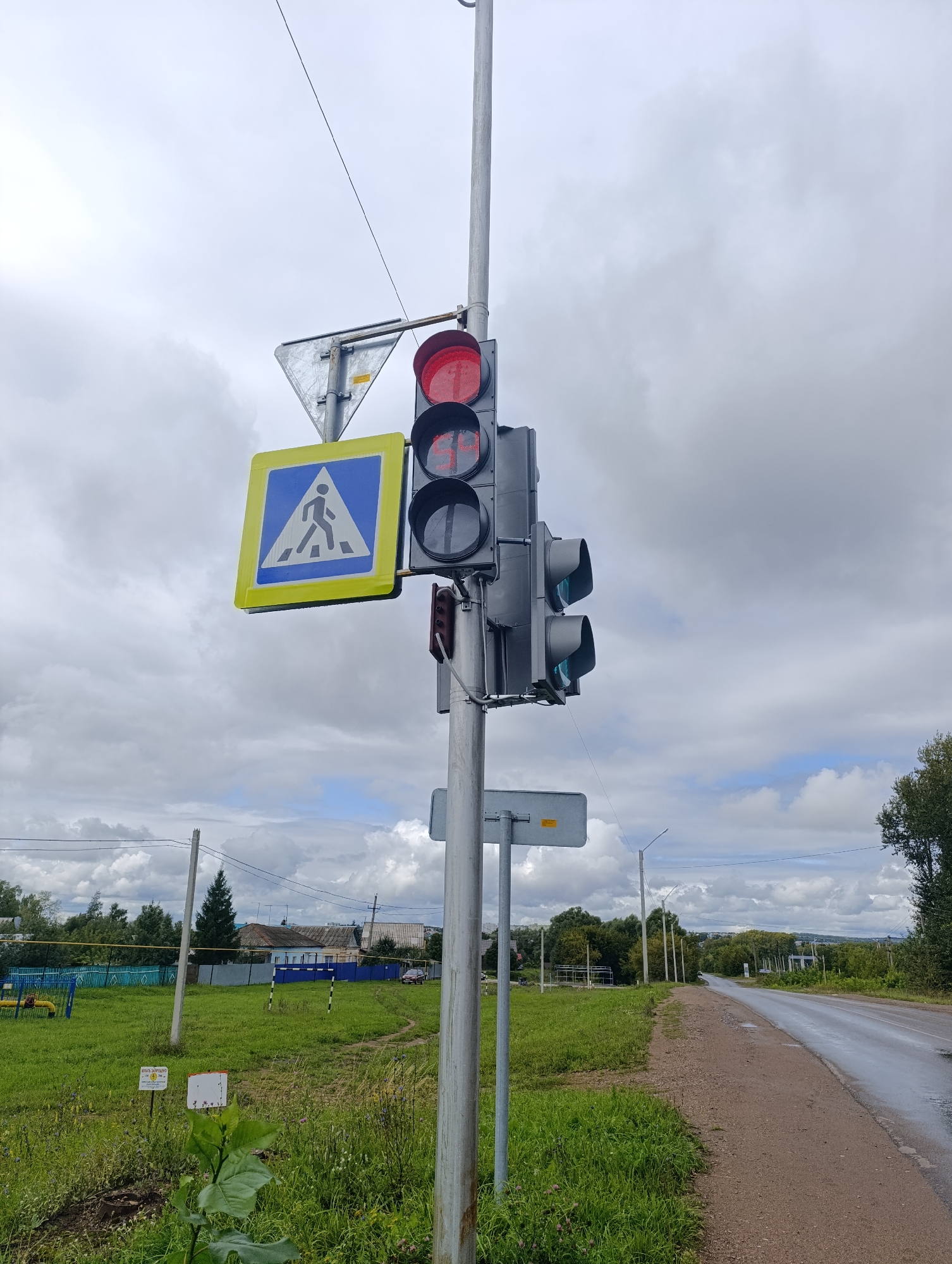 A traffic light with a red signal illuminated hangs on a metal pole beside a pedestrian crossing sign. The pedestrian crossing sign is a blue square with a white triangle inside, depicting a walking figure. The traffic light also has a pedestrian signal with a red illuminated walking figure. In the background, a road stretches into the distance, with a green grassy area to the left and trees and houses on the right. The sky is overcast with clouds. The scene appears to be an intersection in a rural or suburban area.
