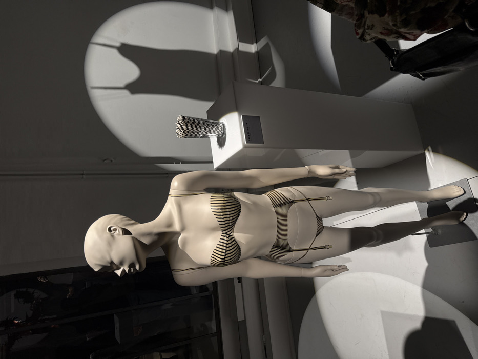 The image depicts an indoor exhibition space or luxury retail environment in Paris, France, illuminated by dramatic spotlights. The central subject is a full-body, light beige, bald female mannequin, posed with its head turned to the left. The mannequin wears a two-piece lingerie set featuring horizontal black stripes on a beige background: a bandeau-style bra with thin shoulder straps and a high-waisted garter belt brief with four descending black garters terminating in metallic clips. A thin, light-colored bracelet is visible on the mannequin's right wrist. The mannequin stands on a dark, rectangular metal base beneath its left foot.
To the right of the mannequin, a white rectangular pedestal holds a clear glass vase containing numerous upright black and white diagonally striped straws or rods. Below this, a small dark plaque on the pedestal's front face reads "L'AS DE MATIERE." The background consists of plain light-colored walls. Intense spotlights cast circular pools of light and strong shadows across the scene. Notably, a distinct shadow of a camisole or slip with spaghetti straps is projected onto the wall behind the pedestal. In the lower right corner, a dark object with a floral pattern, possibly a bag or clothing, is partially visible, suggesting the presence of an observer. The upper left background shows indistinct dark reflections in what appears to be a large window or mirrored surface.