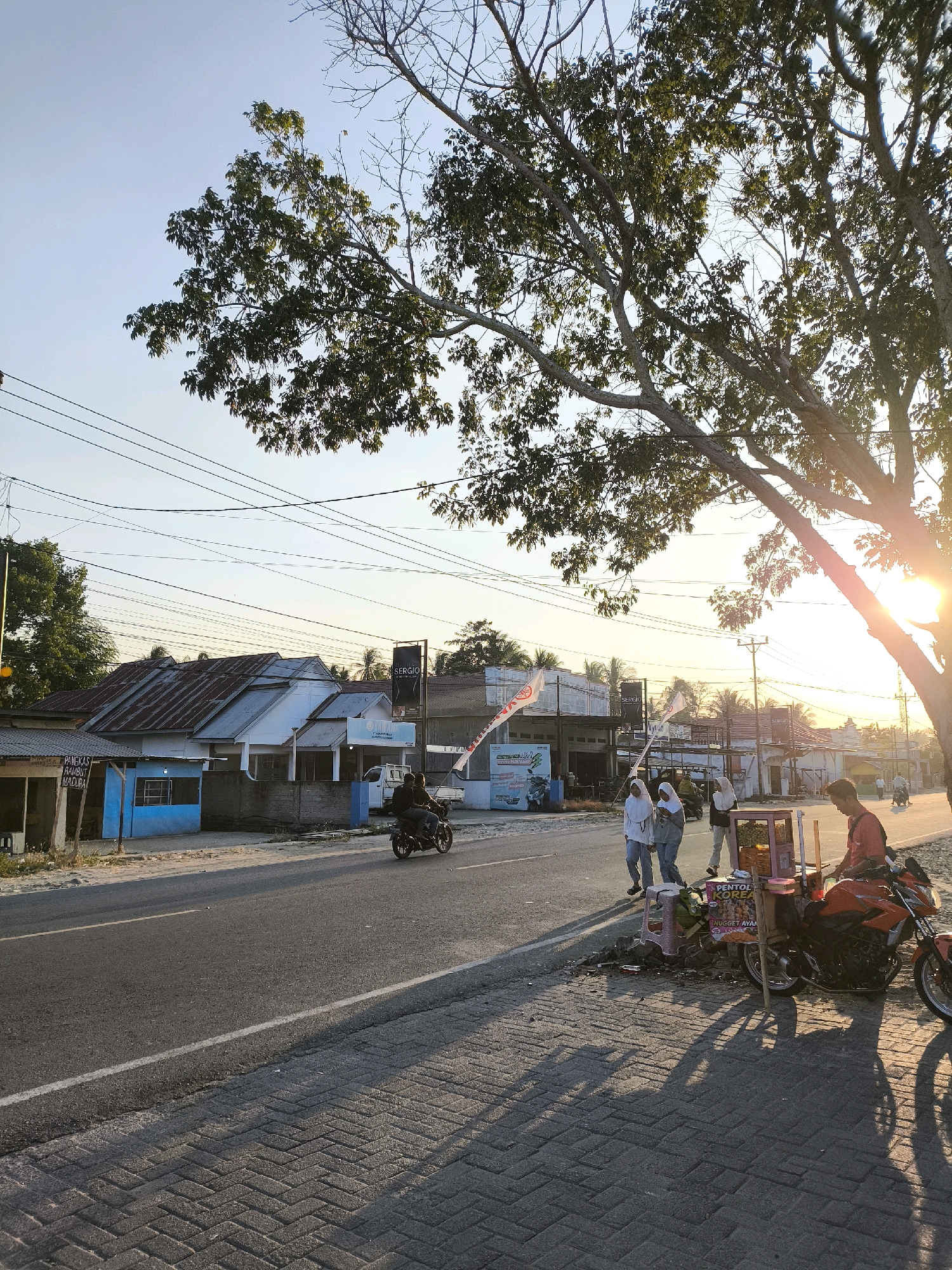 This is a street scene in Tenilo, Indonesia, captured during the golden hour. The sun is low in the sky, casting long shadows across the paved sidewalk and asphalt road. On the left side of the road, there are several buildings with corrugated metal roofs, some painted blue and white. A motorcycle is being ridden on the road. On the right side of the road, a person is sitting on an orange motorcycle next to a food cart. Three young women in hijabs are walking on the sidewalk. A large tree with sparse leaves casts a significant shadow over the scene. Utility lines are visible strung across the sky. The overall atmosphere is that of a quiet, late afternoon in a semi-urban environment.