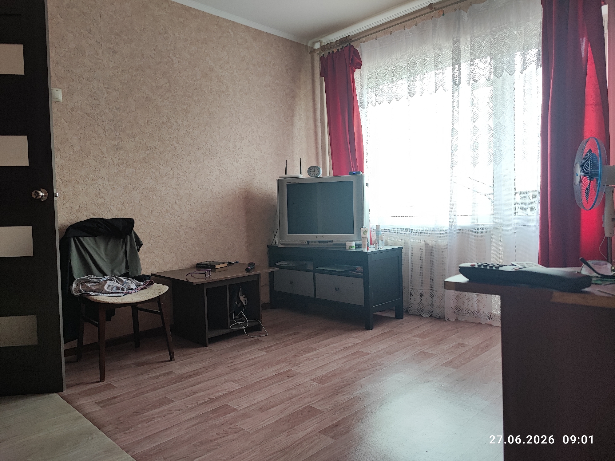 The image captures an interior view of a room, likely a living area, located in Lytkarino, Russia. The room features a light-colored textured wallpaper on the walls and a wooden laminate floor. On the left side of the image, a dark door with lighter rectangular panels is visible. To the right of the door, a simple wooden chair is positioned, draped with a dark jacket and a patterned piece of clothing. Adjacent to the chair is a small, dark wooden table with a book and glasses placed on top, and a pair of headphones with a cord hanging below.
Further to the right, a television set rests on a dark entertainment unit, which has two drawers and open shelving. A router with antennas is placed behind the television, and a small analog clock is also visible on top of the TV. A pair of red curtains and white lace curtains are drawn across a large window that takes up a significant portion of the right side of the room, allowing natural light to illuminate the space. A white electric fan is positioned to the right of the red curtains. In the foreground on the right, a corner of a dark brown table is visible, with a remote control resting on its surface. The timestamp "27.06.2026 09:01" is superimposed on the bottom right of the image. There are no people present in the scene.