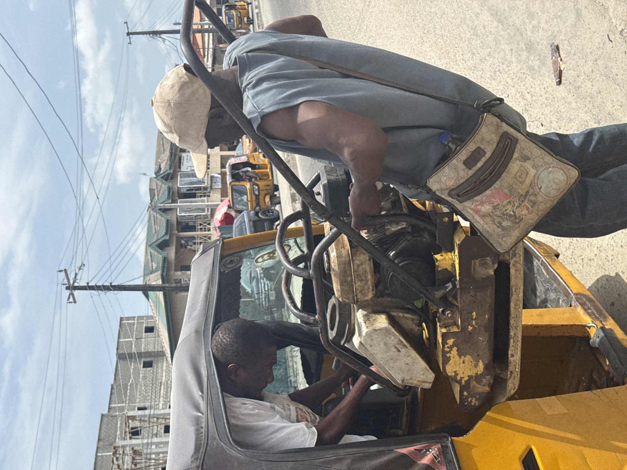 Two men are inspecting the engine of a yellow motorized tricycle, also known as a "Keke," on a street in Ado, Nigeria. One man, wearing a tan baseball cap, a light blue shirt, and jeans with a patterned messenger bag, is leaning over the engine. The second man is sitting inside the Keke, wearing a white shirt, and examining the engine from within the vehicle. The surroundings include buildings and utility poles with wires. Other yellow Kekes and vehicles are visible in the background, parked along the street. A piece of debris is on the road near the Keke.