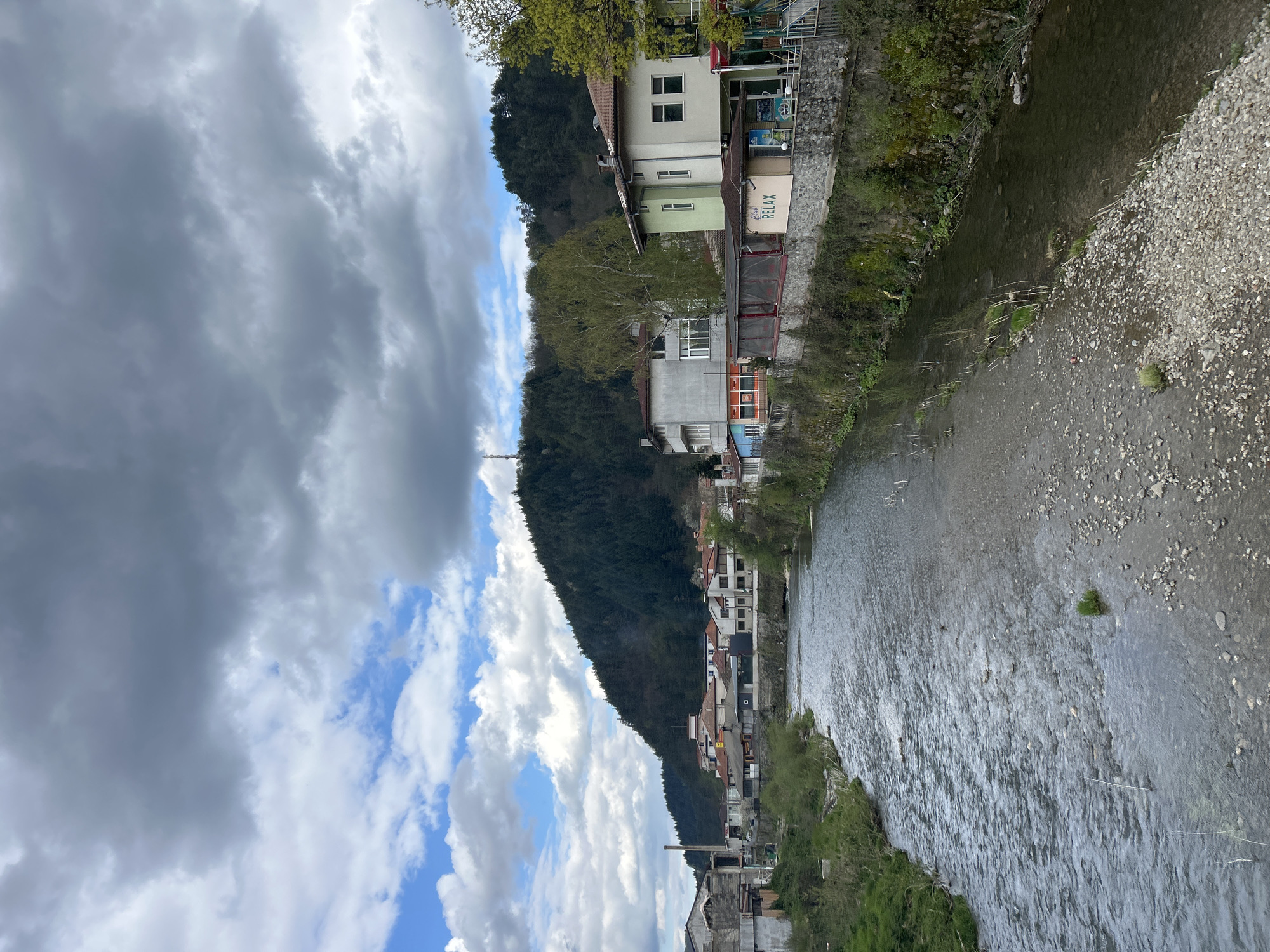 This media file captures a picturesque view of a river flowing through a valley in Zlatograd, Bulgaria. The image, originally rotated, shows a dynamic sky overhead, featuring a mix of dark, heavy clouds and patches of bright blue sky with white cumulus clouds, suggesting a day with variable weather.
The river, a prominent subject, occupies the middle and foreground of the scene. Its water appears somewhat turbulent with visible ripples and reflections, and its bed is clearly visible in shallower areas, characterized by numerous pebbles and small rocks. Sparse green vegetation, including grass and small bushes, grows along the immediate banks of the river.
Along the left bank of the river, a line of buildings stands, integrated into the landscape. These structures appear to be a mix of residential houses and possibly commercial establishments, displaying various architectural styles and colors, including light green, white, and grey facades. Many have balconies and windows facing the river. On one of these buildings, a sign clearly reads "Cafe RELAX".
Rising steeply behind the buildings and dominating the right side of the scene is a densely forested mountain or hillside. The trees appear dark green, indicating a thick covering of coniferous or deciduous trees. The overall setting depicts a small town nestled between a waterway and mountainous terrain, giving a sense of a peaceful, natural environment intertwined with human settlement. The time of day appears to be daytime, likely during daylight hours given the ambient light. No specific human activity is discernible.