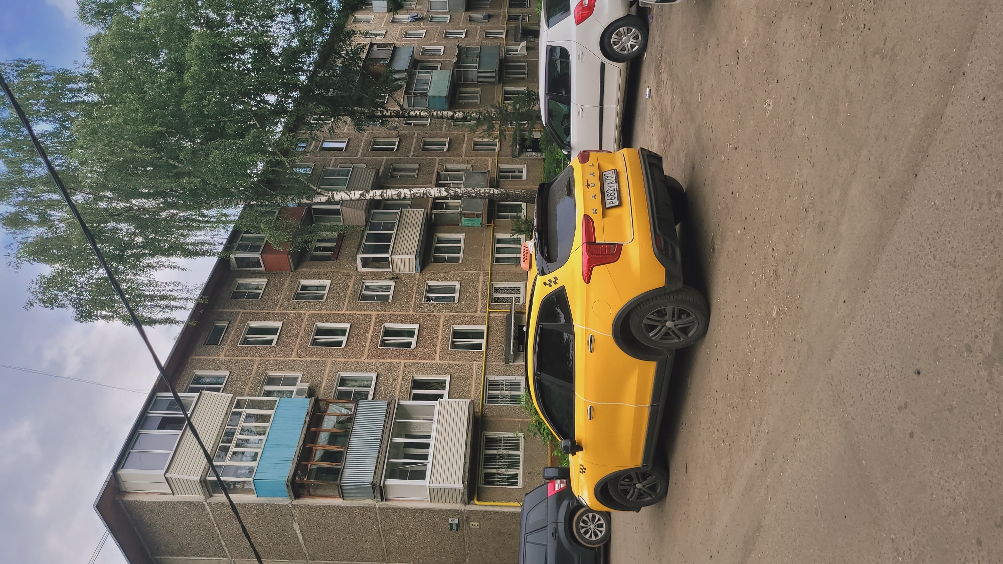 An eye-level, exterior, full shot shows a yellow taxi parked next to a concrete apartment building. The taxi, a Kia Rio, is facing right and is oriented vertically in the frame. It has a black spoiler on top and the license plate reads "P682AX | 97". A taxi sign is visible on the roof of the car. A white car is parked in front of the taxi, also oriented vertically. The apartment building is tall and grey, with many windows and balconies. A large tree with green leaves is growing in front of the building. The ground is paved with asphalt. The sky is cloudy. The scene appears to be a residential area, possibly in Russia, given the license plate. No people are visible in the frame. The lighting suggests it is daytime, possibly overcast.