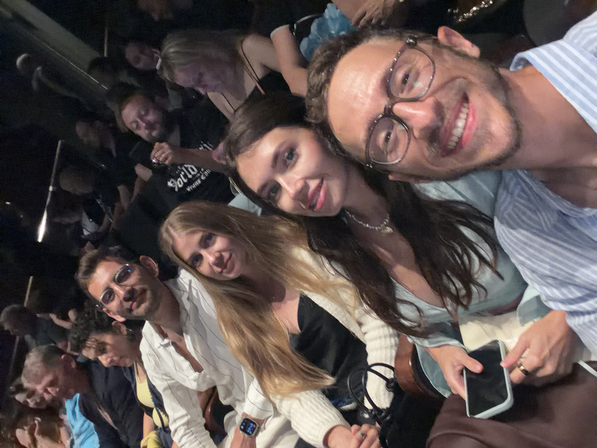 In an indoor, dimly lit environment, likely a theatre or concert hall in Tel Aviv, Israel, a group of people are seated, attending an event. In the foreground, a man with glasses and a light blue and white striped shirt smiles broadly at the camera, holding a smartphone. To his left, a woman in a light blue top looks towards the camera with a pleasant expression, holding a black phone in her hands. Behind them, other individuals are visible, including a woman with long blonde hair wearing a white cardigan over a black top, and a man with short dark hair and glasses in a white striped shirt. The audience is seated in rows, suggested by the arrangement and the dark background. The atmosphere appears relaxed and social, with people engaged in taking a selfie.