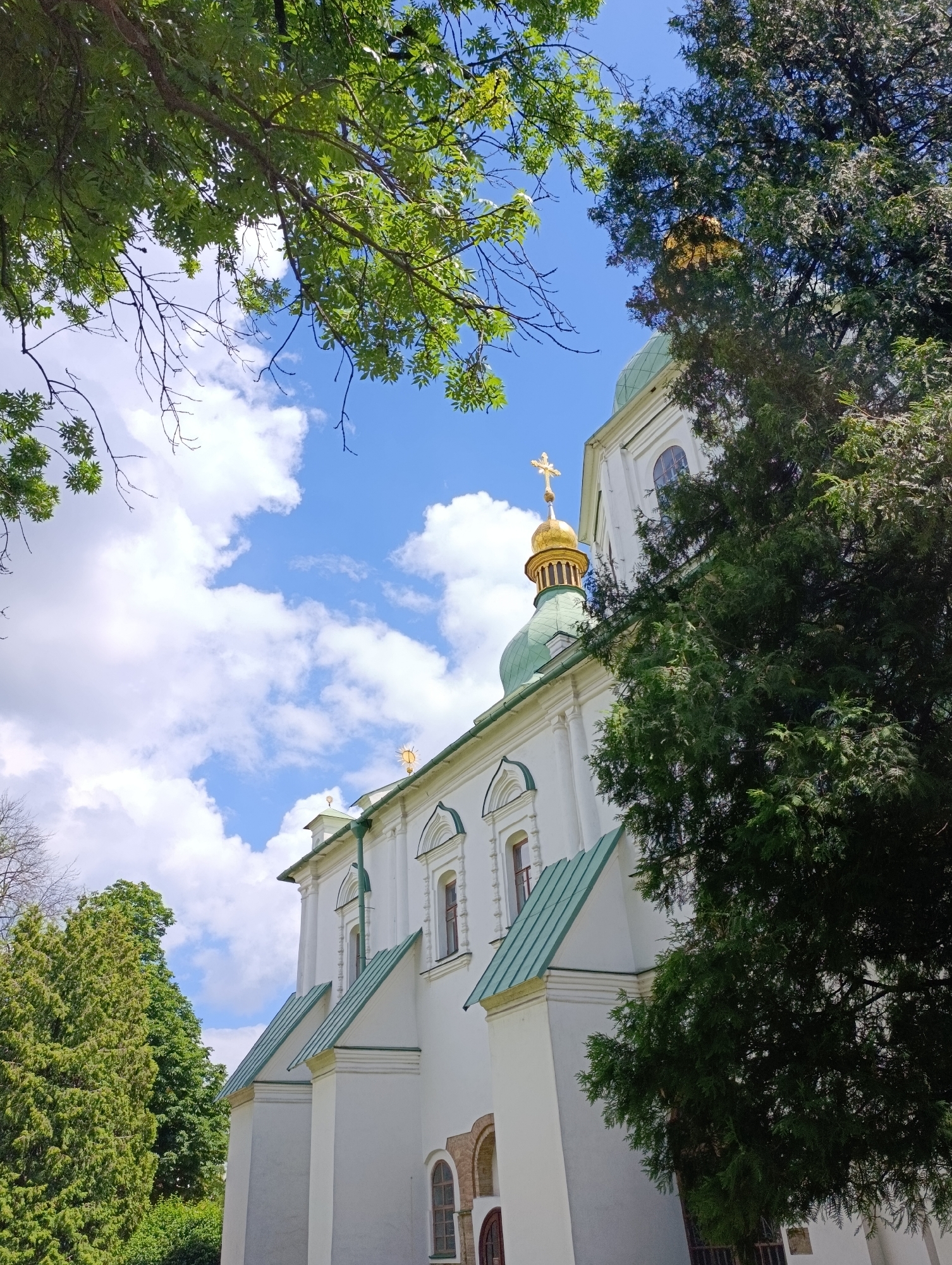 This is a low-angle, eye-level outdoor photograph of the St. Sophia's Cathedral in Kyiv, Ukraine. The white facade of the cathedral, with its green roof and golden dome topped with a cross, is partially obscured by lush green trees. The sky above is a bright blue with scattered white clouds. The image composition focuses on the architectural details of the cathedral and the surrounding natural elements.