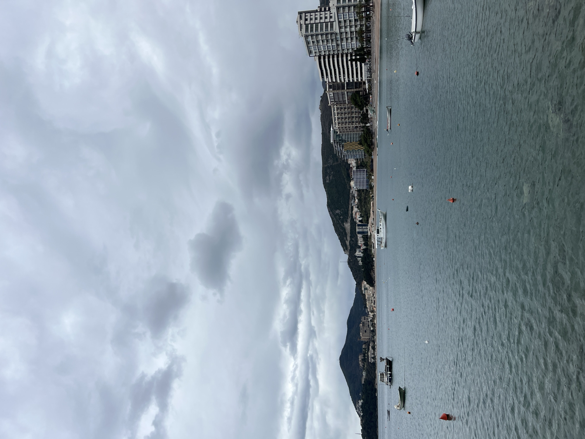 The image depicts a coastal cityscape, likely Budva, Montenegro, under an overcast sky. Buildings of various sizes, including high-rise structures, line the coastline at the base of tree-covered hills. Several boats, ranging from small vessels to larger yachts, are scattered across the water. Orange buoys mark the water's surface. The sky is dominated by heavy, grey clouds.