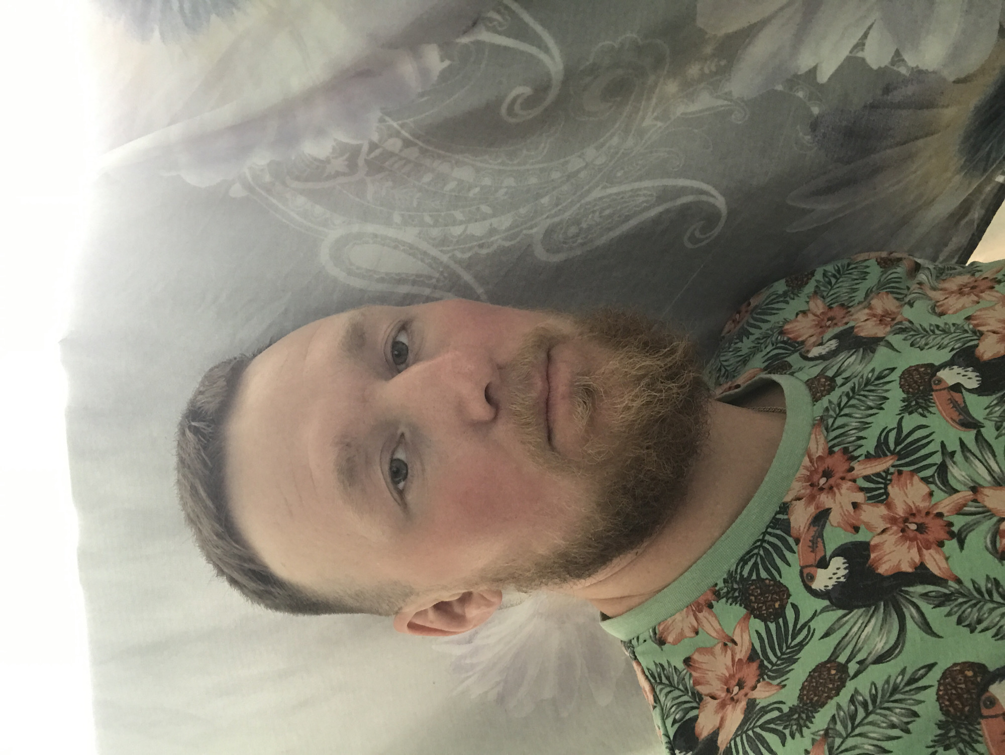 The image shows a man with fair skin, short, light brown hair, and a light brown beard. He is wearing a green t-shirt with a floral and toucan pattern. He appears to be lying down, possibly on a bed, as patterned bedding is visible in the background. The image appears to be taken indoors. The city is Unknown, Unknown.